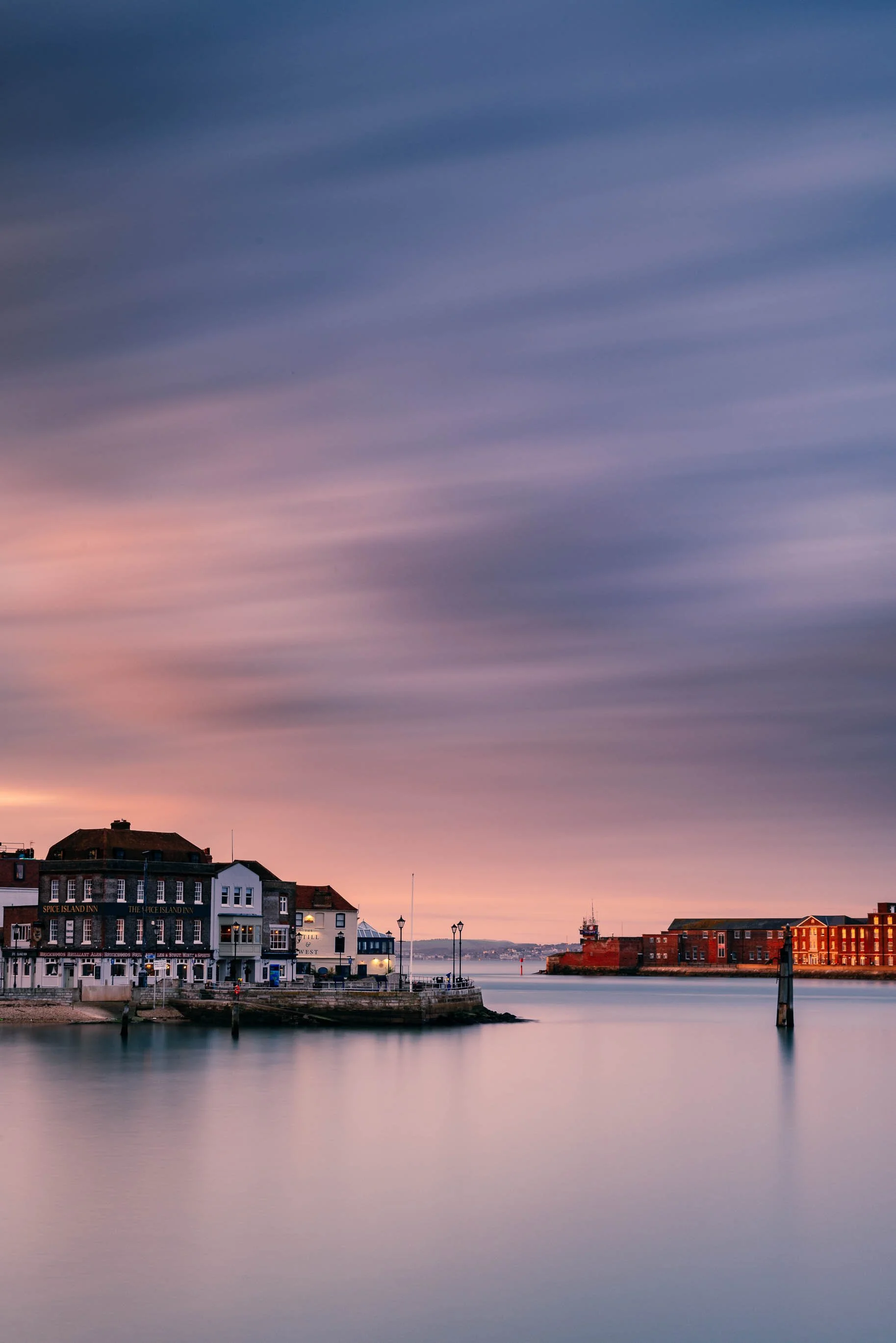 A calm harbor at sunset with pastel-colored skies, historic buildings along the waterfront, and reflections on the water.