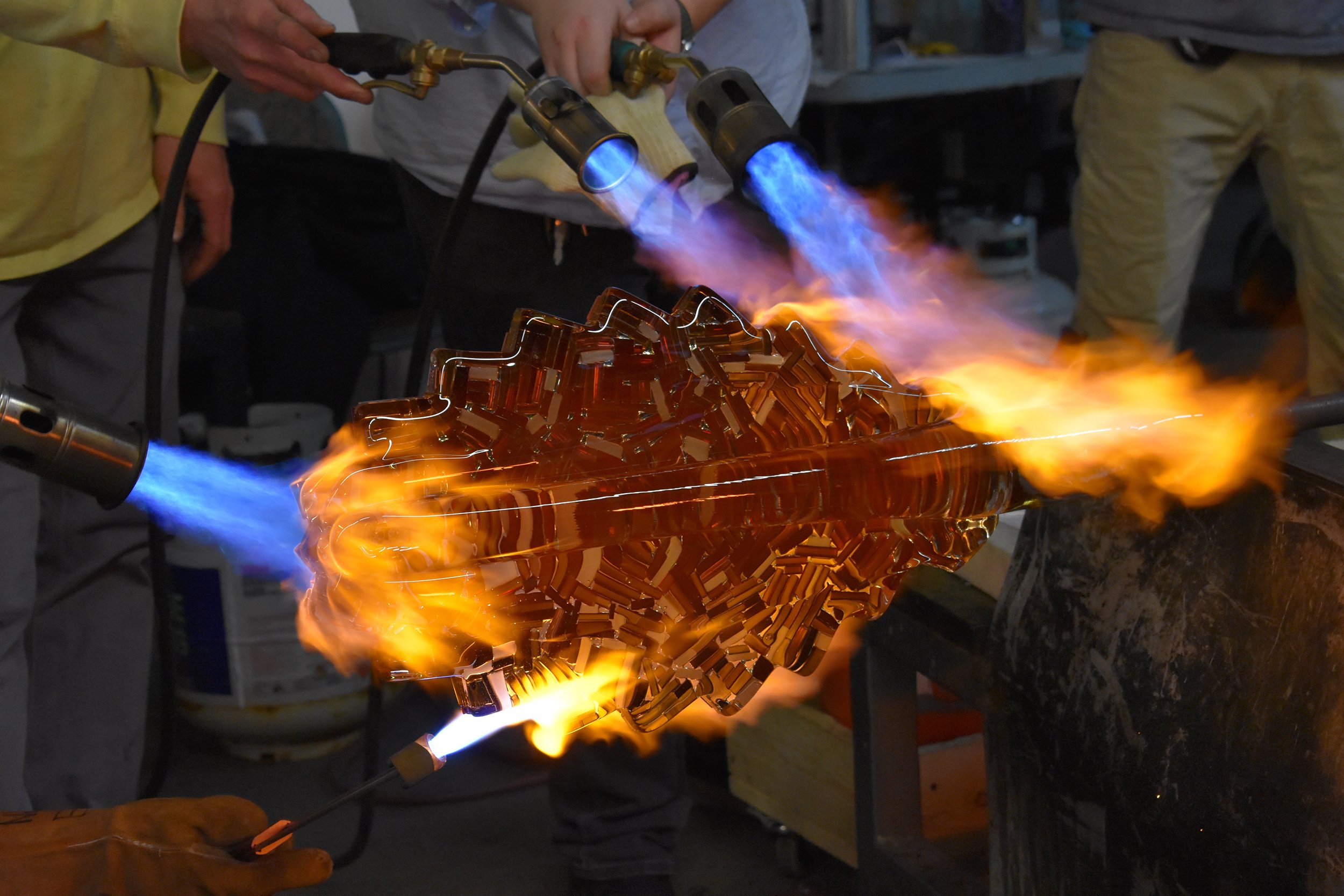 Multiple torches being used to maintain heat on a large glass sculpture during a demonstration for students.