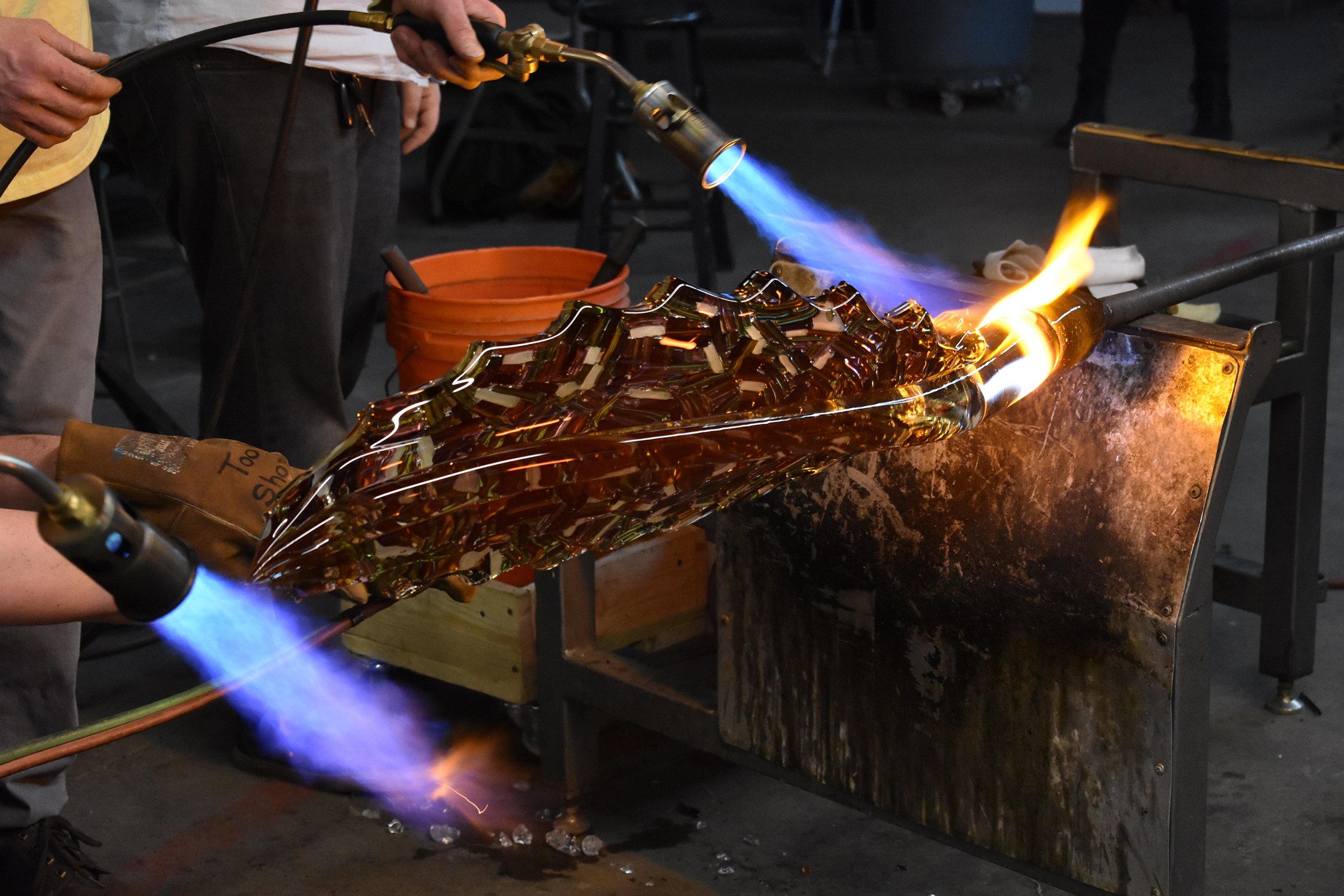 Multiple torches being used to maintain heat on a large glass sculpture during a demonstration for students.