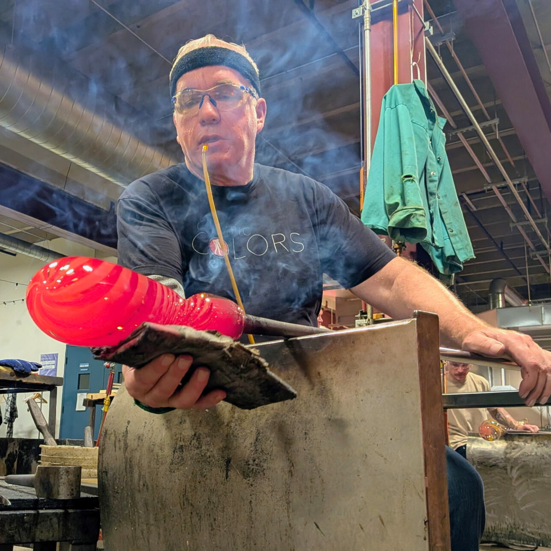 A glassblower wearing safety glasses and a black T-shirt, holding a red molten glass piece with tools, working at a furnace in a studio.