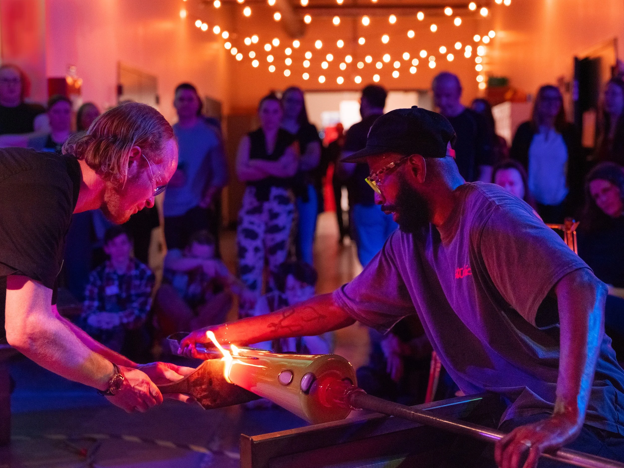 Two glassblowers are working together to create a glass object, with an audience observing in the background. The scene is lit with warm, colorful lights.
