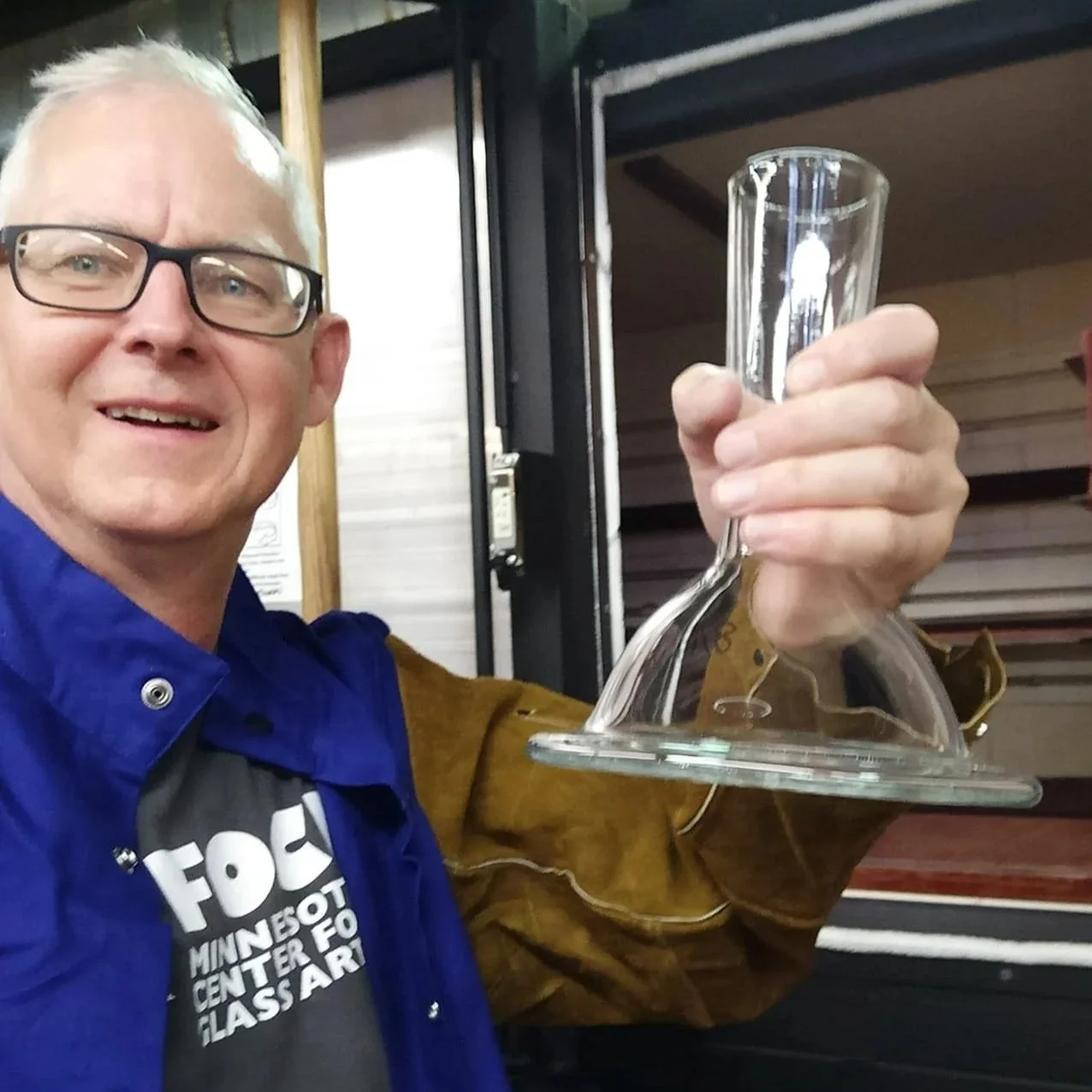 A man smiling, wearing glasses and a blue jacket, holding a glass object with a flat base and a tall, narrow top in front of a window or display case.