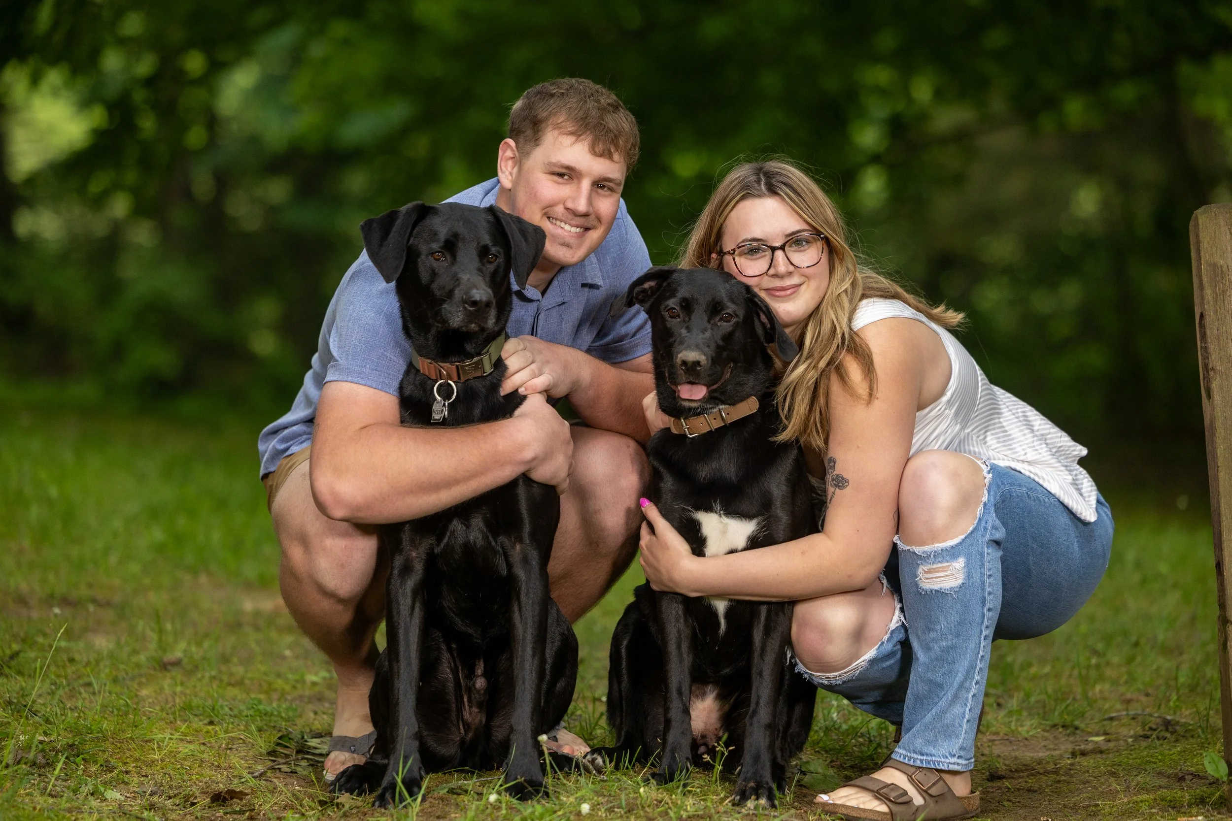 A young man and woman outdoors with two black Labrador dogs, smiling and hugging the dogs, in a green, wooded area.