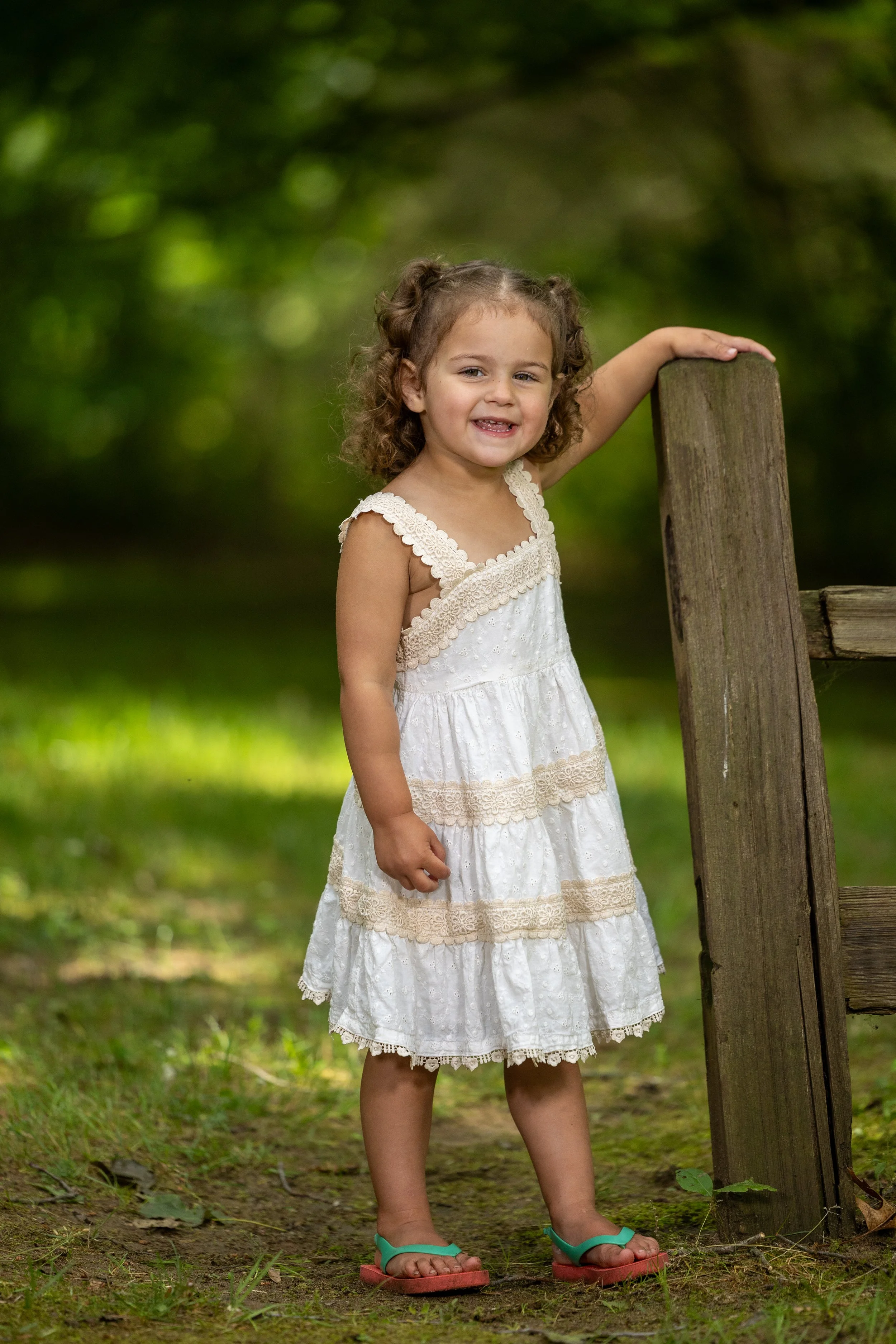 A young girl with curly hair wearing a white, lacy summer dress standing outdoors in a lush green setting.