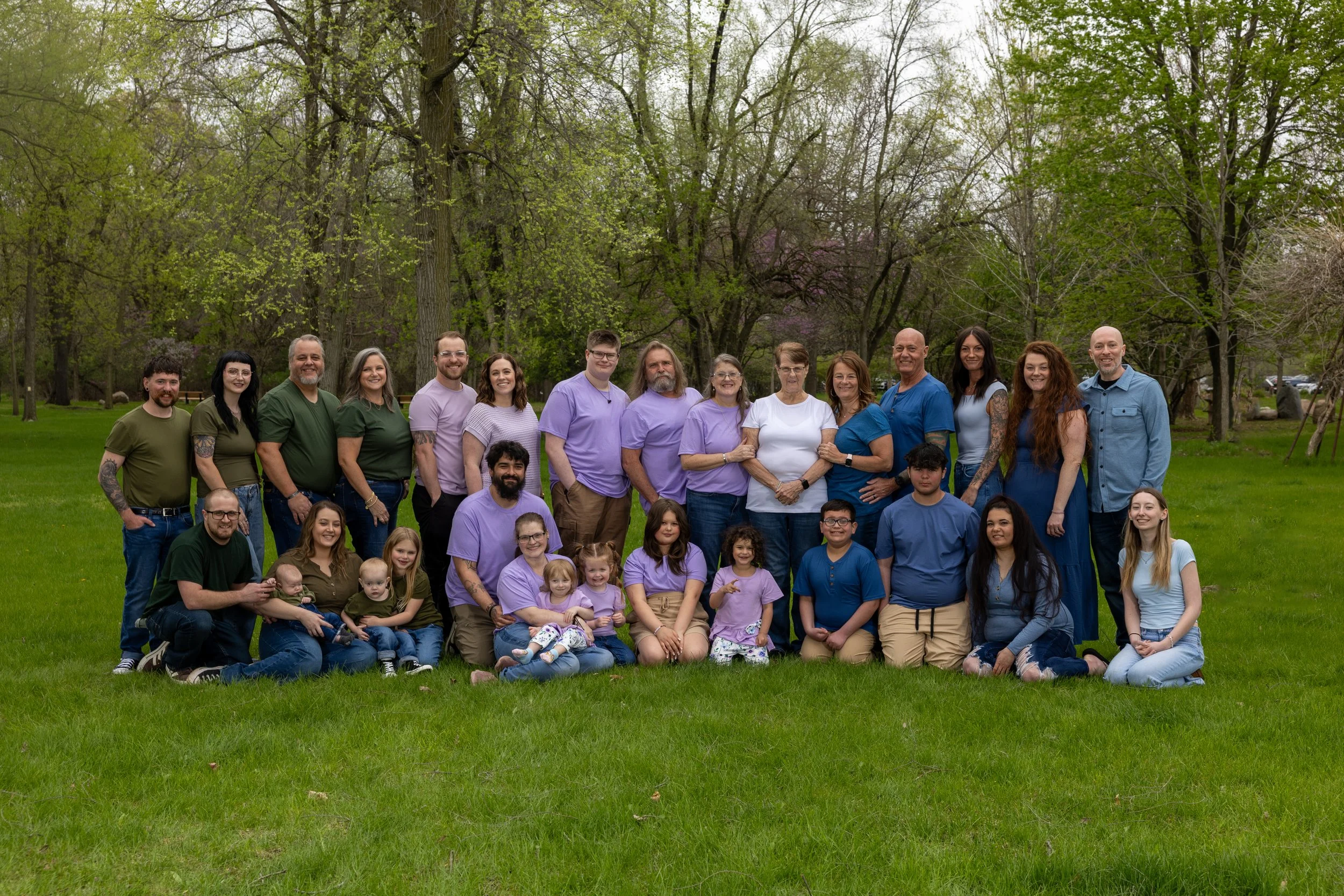 A large group of people, including adults and children, gathered outdoors on a grassy field with trees in the background, smiling for a group photo.
