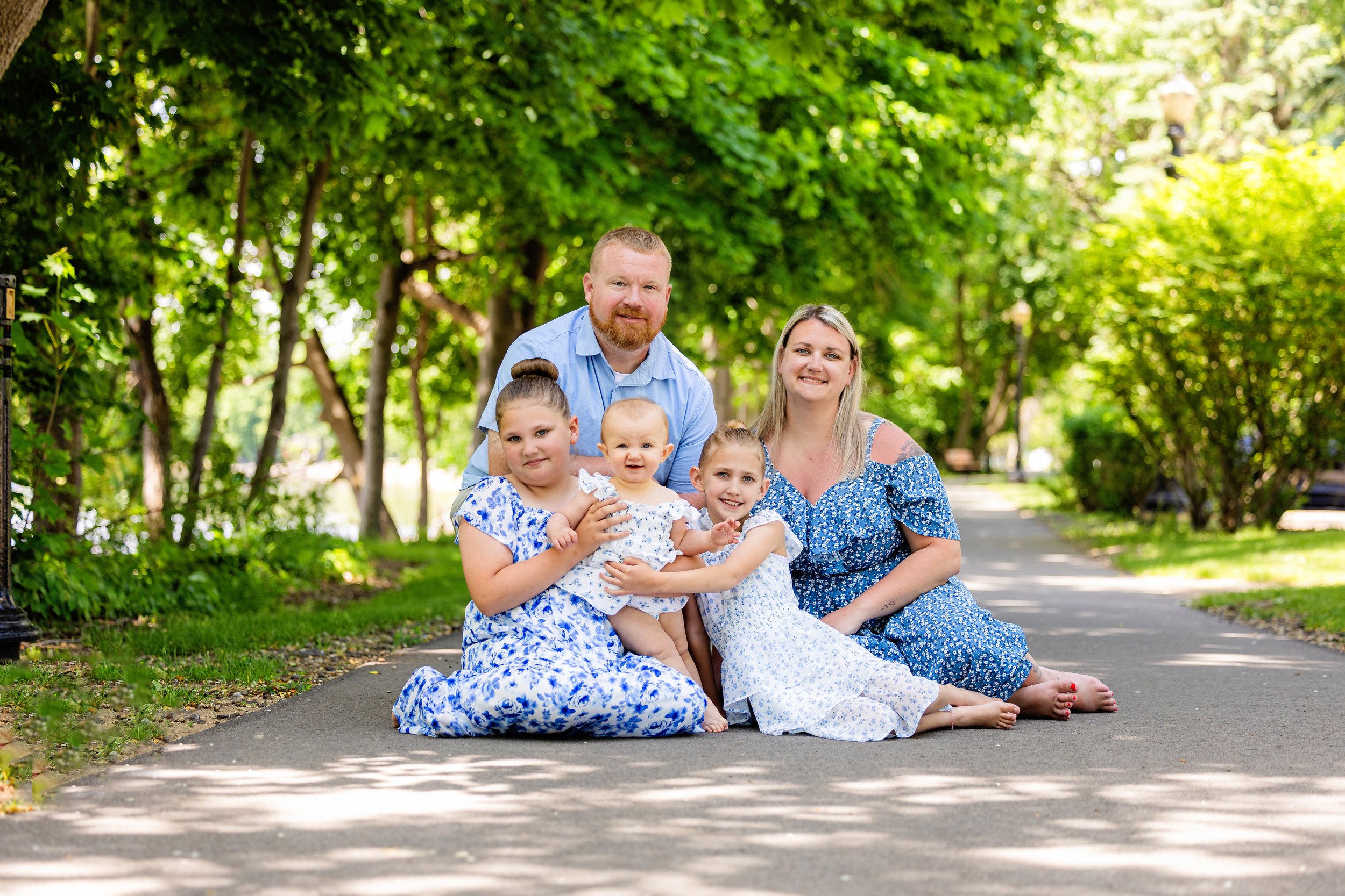 A family of five sitting on the sidewalk in a park, surrounded by green trees, with two adults and three children, all smiling and dressed in blue and white clothing.