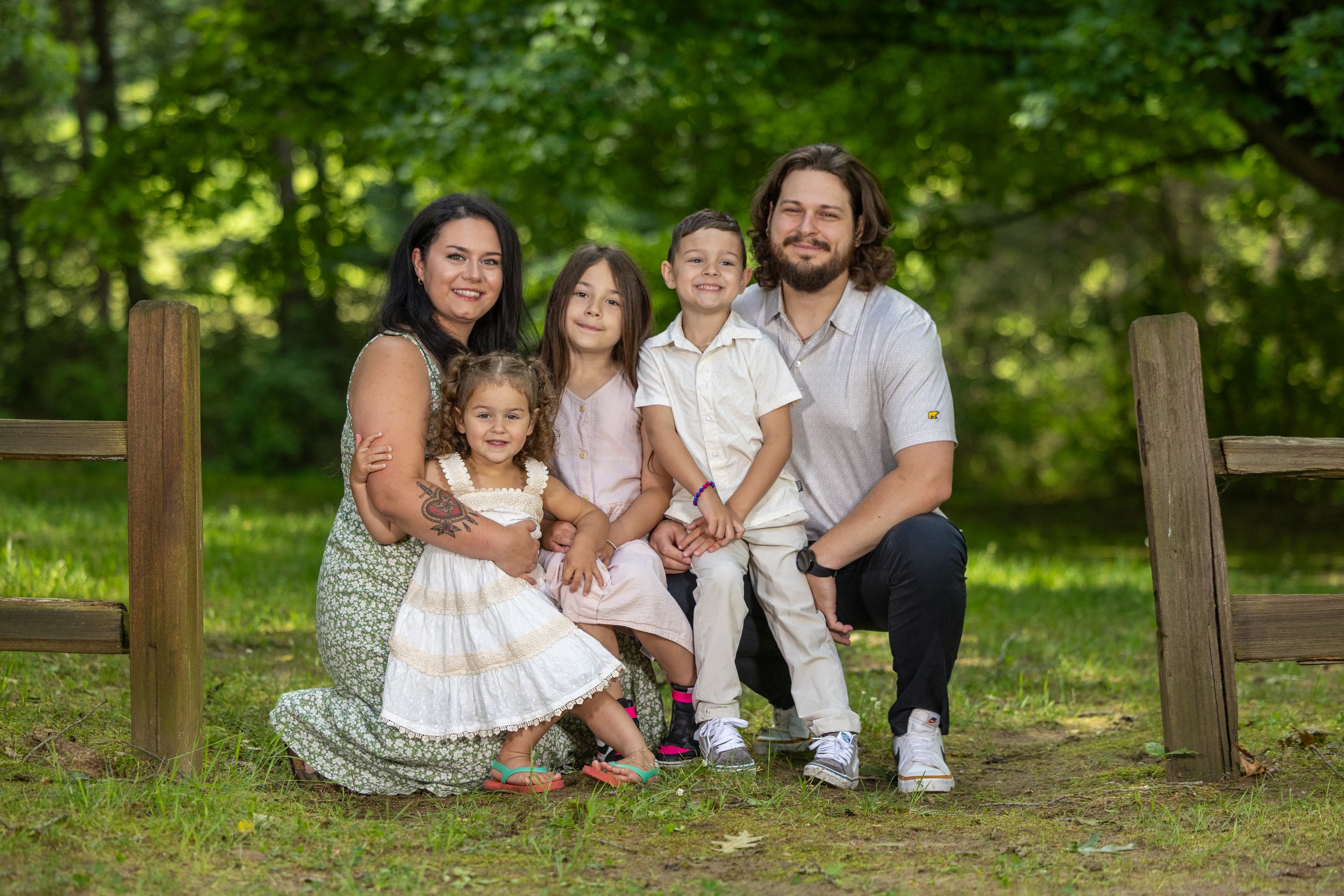 A family of five sitting and kneeling outdoors in a wooded park, smiling at the camera, with a wooden fence behind them and green trees in the background.