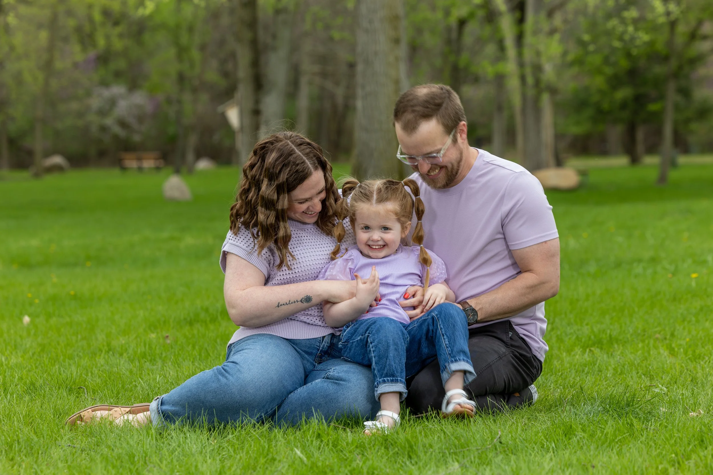 A family of three sitting on the grass in a park, smiling and enjoying each other's company. The mother and father are holding their young daughter, who is smiling at the camera. The background shows trees and a peaceful, outdoor setting.