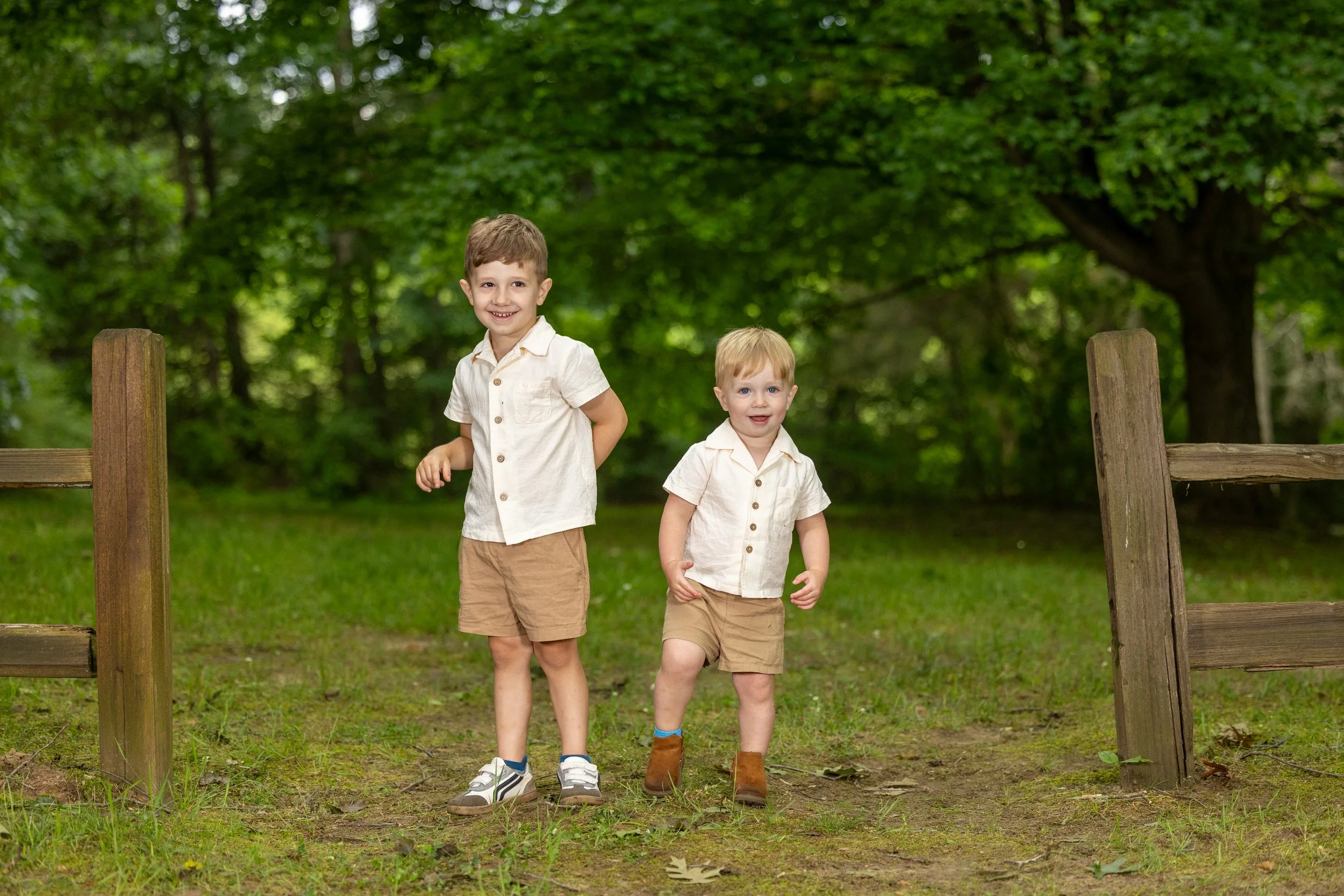Two young boys, one older and taller and another younger, walking outdoors in a park with lush green trees in the background. They are dressed in light colored shirts and khaki shorts, smiling and enjoying a day outside.