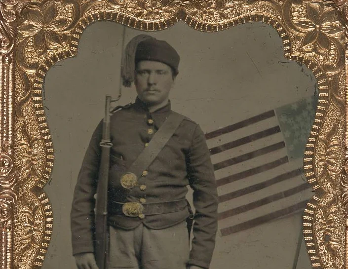 (Unidentified_soldier_in_Union_uniform_with_Zouave_fez_and_bayoneted_musket_in_front_of_painted_backdrop_showing_American_flag)_(LOC)_(5228558775).jpg