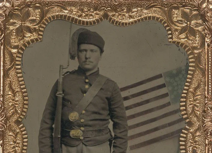 (Unidentified_soldier_in_Union_uniform_with_Zouave_fez_and_bayoneted_musket_in_front_of_painted_backdrop_showing_American_flag)_(LOC)_(5228558775).jpg