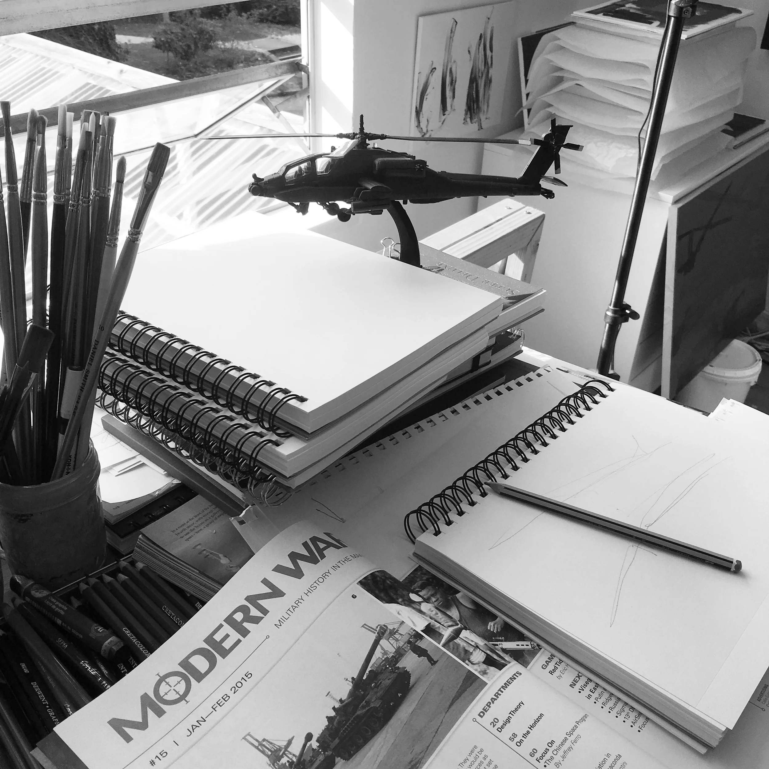 Black and white image of a workspace with sketchbooks, a pencil, a model helicopter, paintbrushes, a military history magazine, and art supplies by a window.
