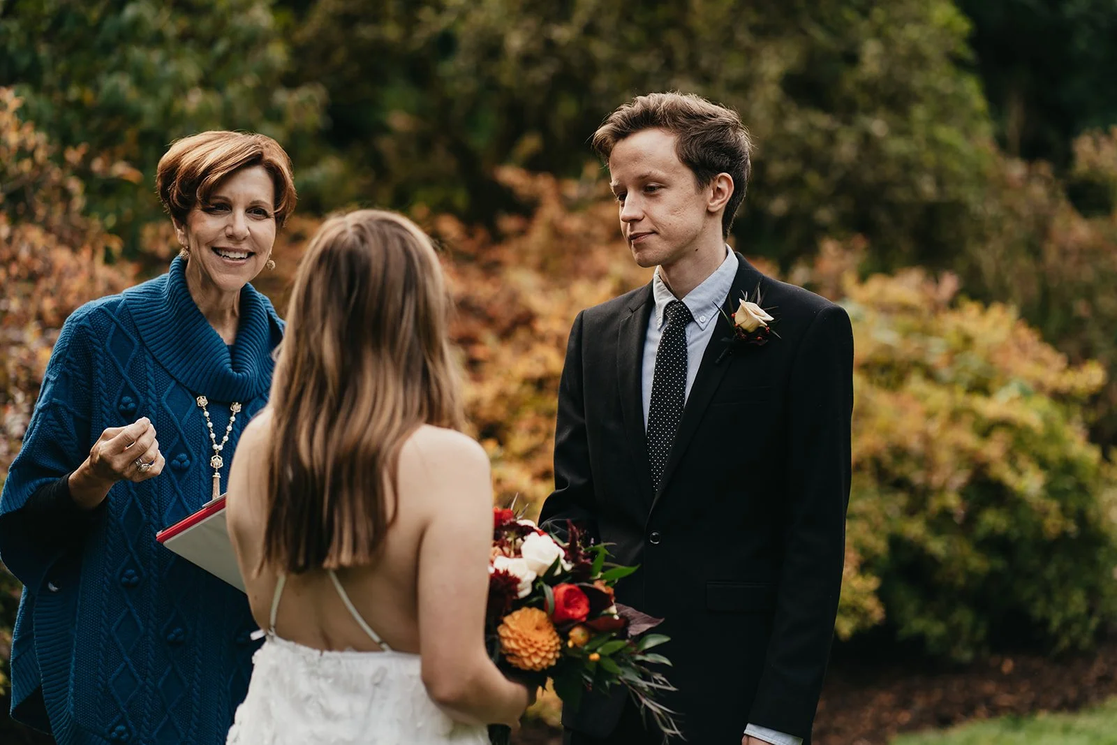 an elopement couple say their vows in front of an officiant in a blue coat with Fall foliage behind them