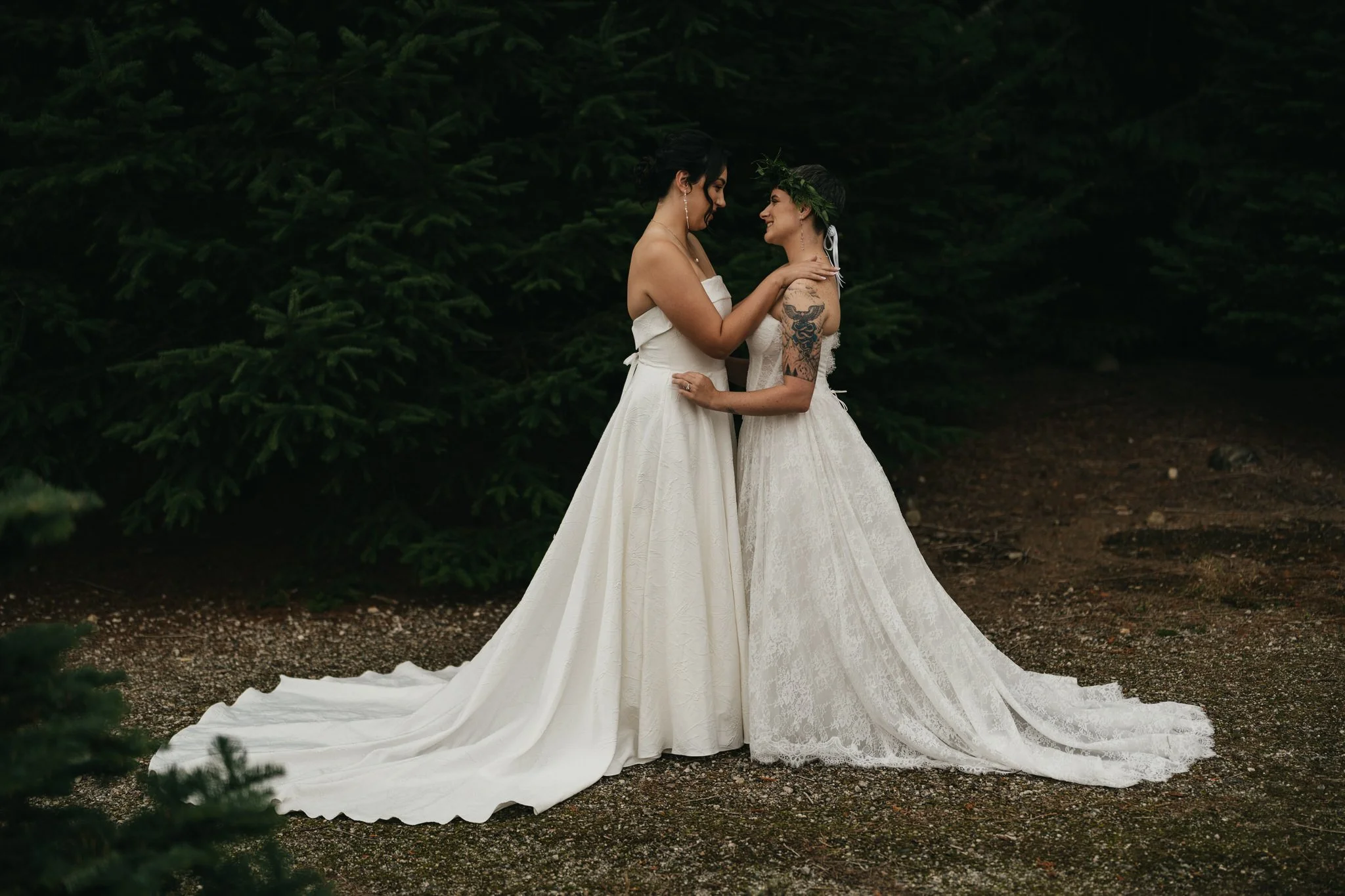two brides in white wedding dresses embrace on their wedding day in front of a pine tree