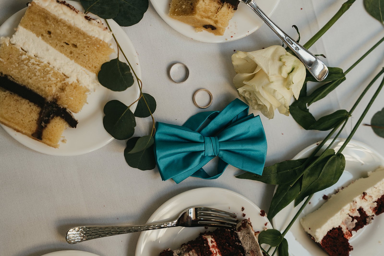 a messy late night flat lay from a wedding with cake, a flower, a grooms bowtie, and their wedding rings