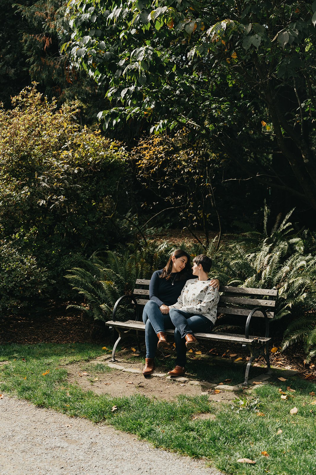 a lesbian couple sits on a bench looking at each other, surrounded by greenery