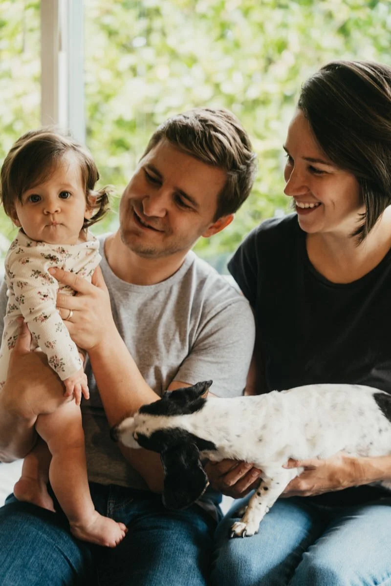 a family of three humans and a dog sit on a couch, the parents are looking at the baby and the baby is looking at the camera