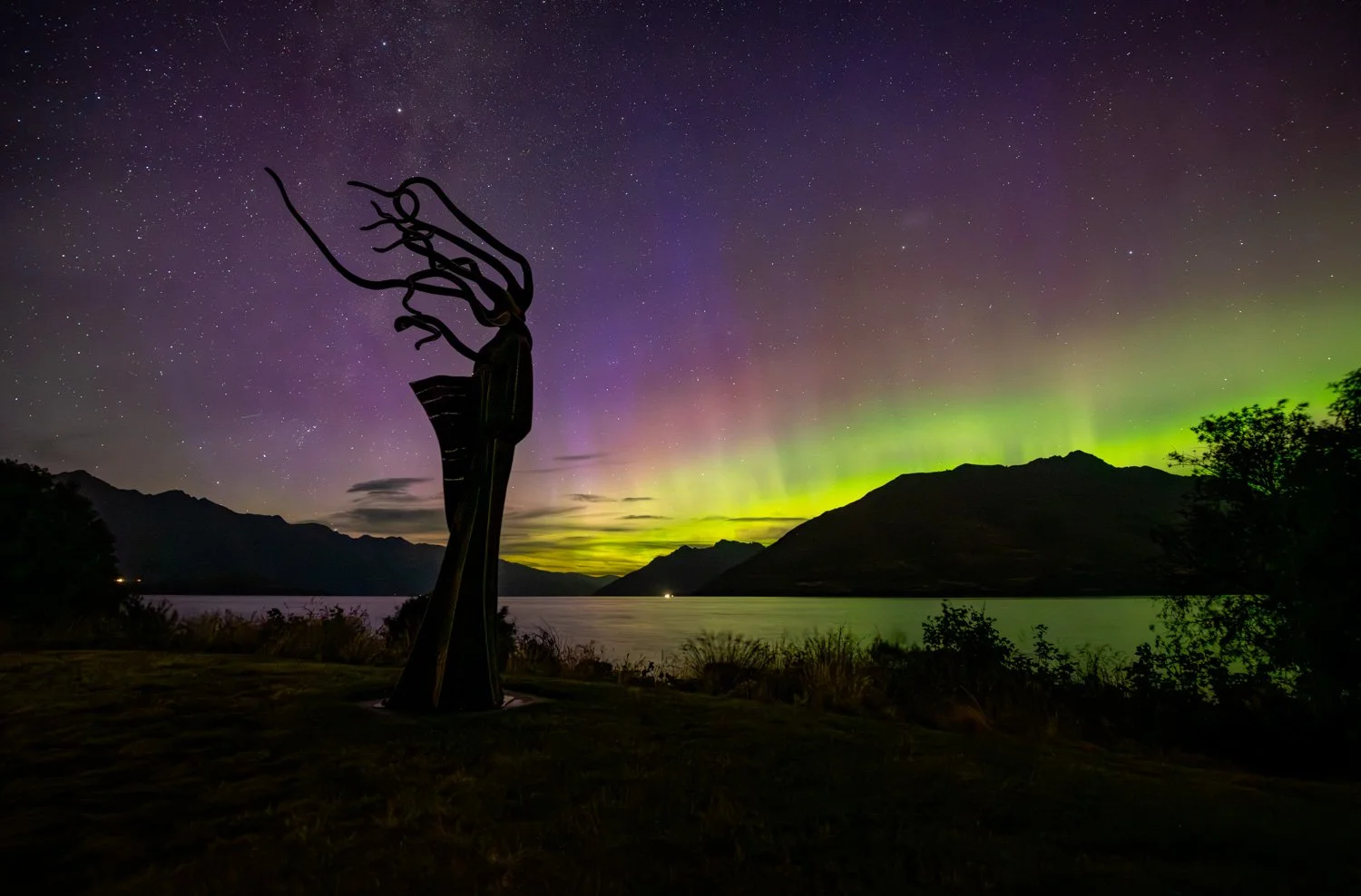 Silhouette of Whakatipu Kiukiu sculpture by Mark Hill against night sky with green and magenta aurora australis, female figure with flowing hair facing across Lake Wakatipu toward the mountains