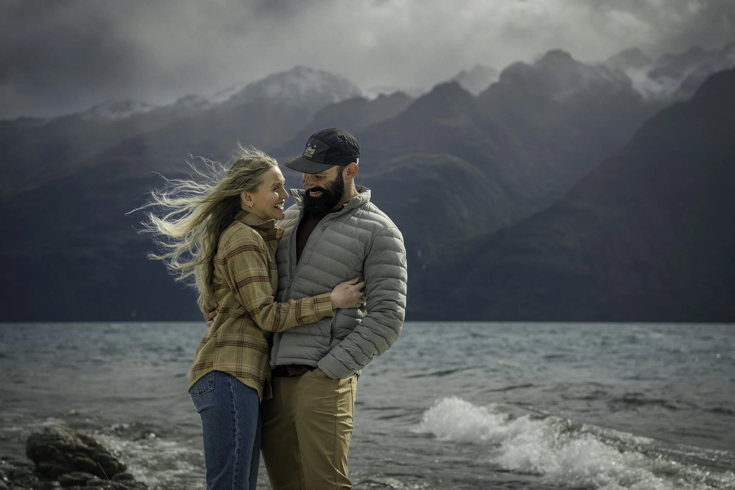 Young honeymooning couple embrace on the shores of Lake Whakatipu as the wind whistles around them, Queenstown, New Zealand