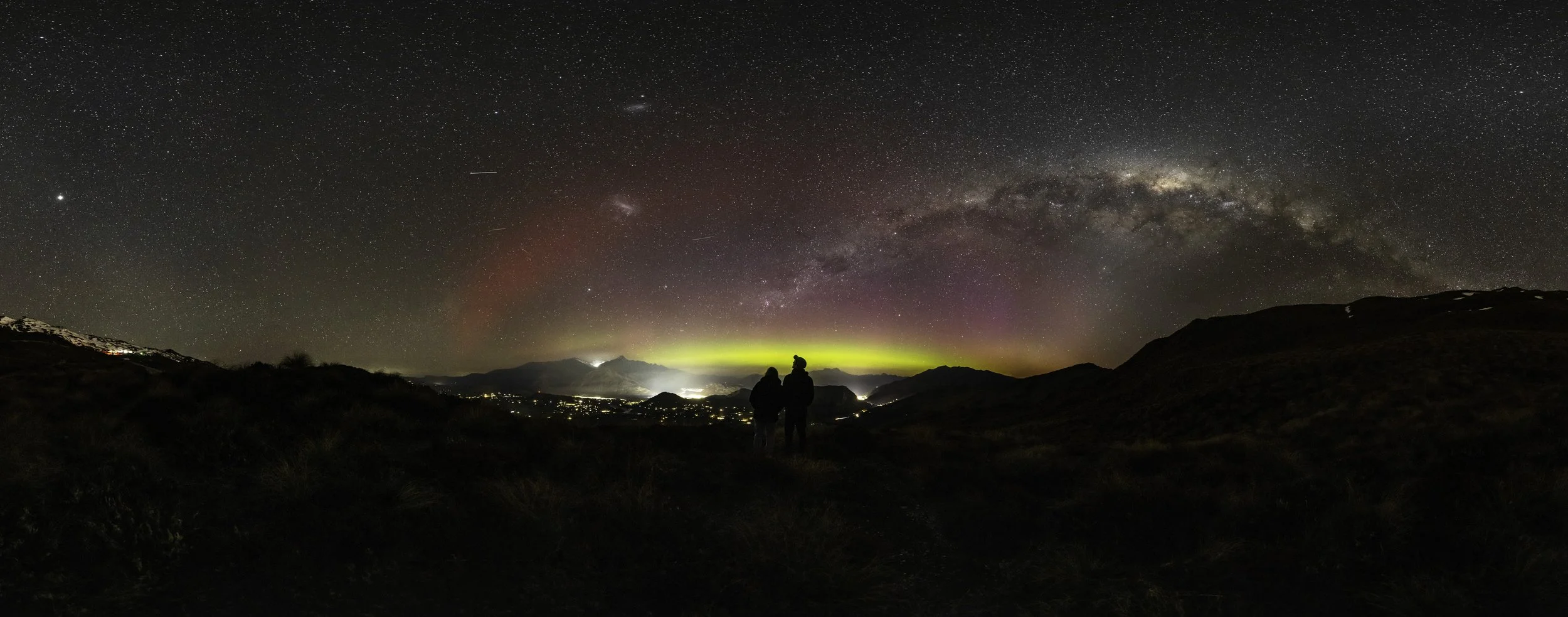 A couple enjoy the view over the Wakatipu Basin beneath a winter Milky Way arch, Aurora Australis and SAR Arc during a Starry Night Adventure, Queenstown, New Zealand