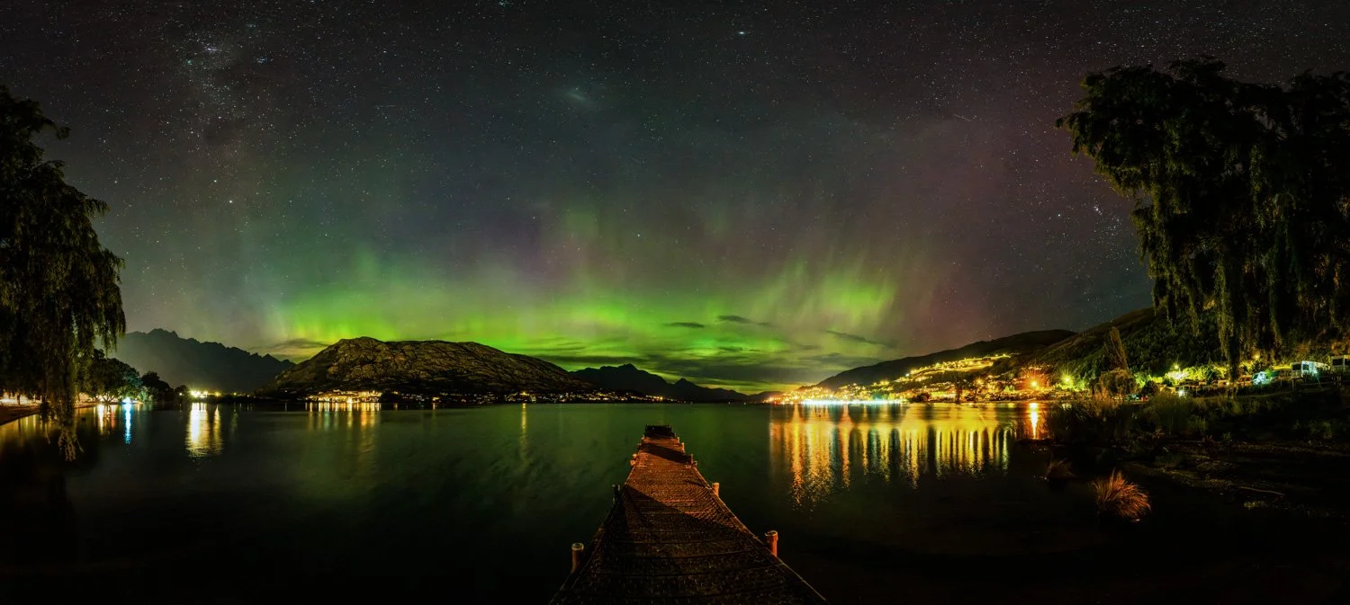 Aurora australis with green beams visible above mountains from Frankton pier, marina and foreground heavily lit by orange sodium street lights creating strong light trespass