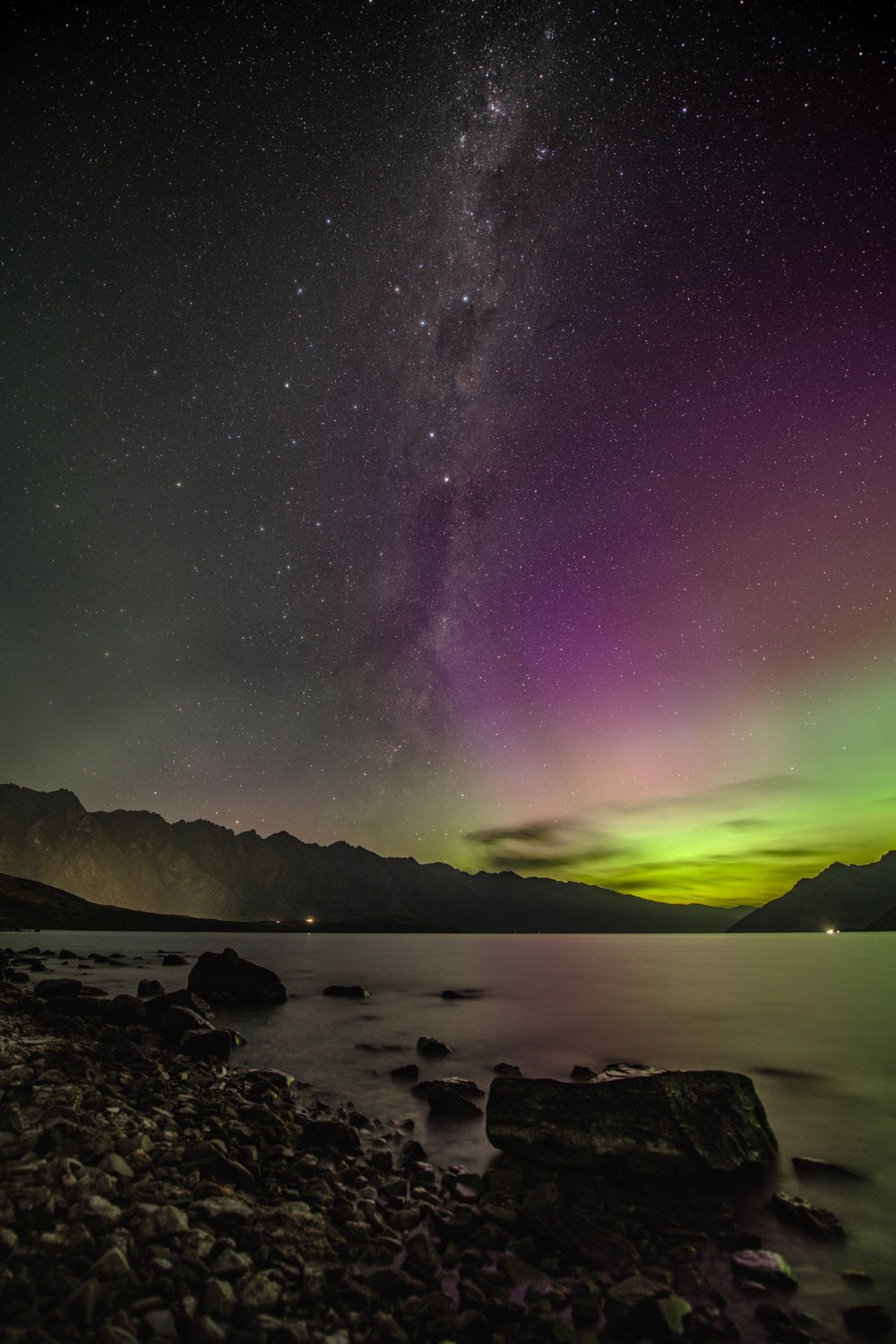Milky Way arcing across dark night sky above Lake Wakatipu and the Remarkables mountain range, Coalsack Nebula and Southern Cross visible, faint green aurora glow on horizon