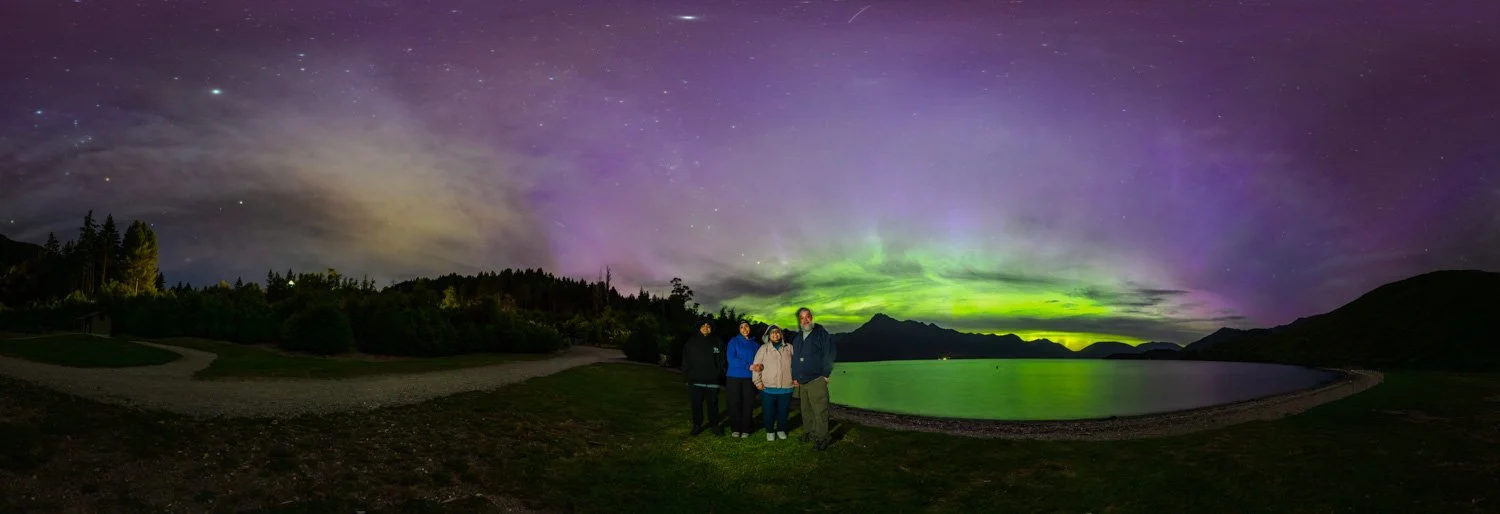Family of four standing on shore of Lake Wakatipu illuminated against vibrant green and magenta aurora australis filling the sky above the Remarkables mountain range