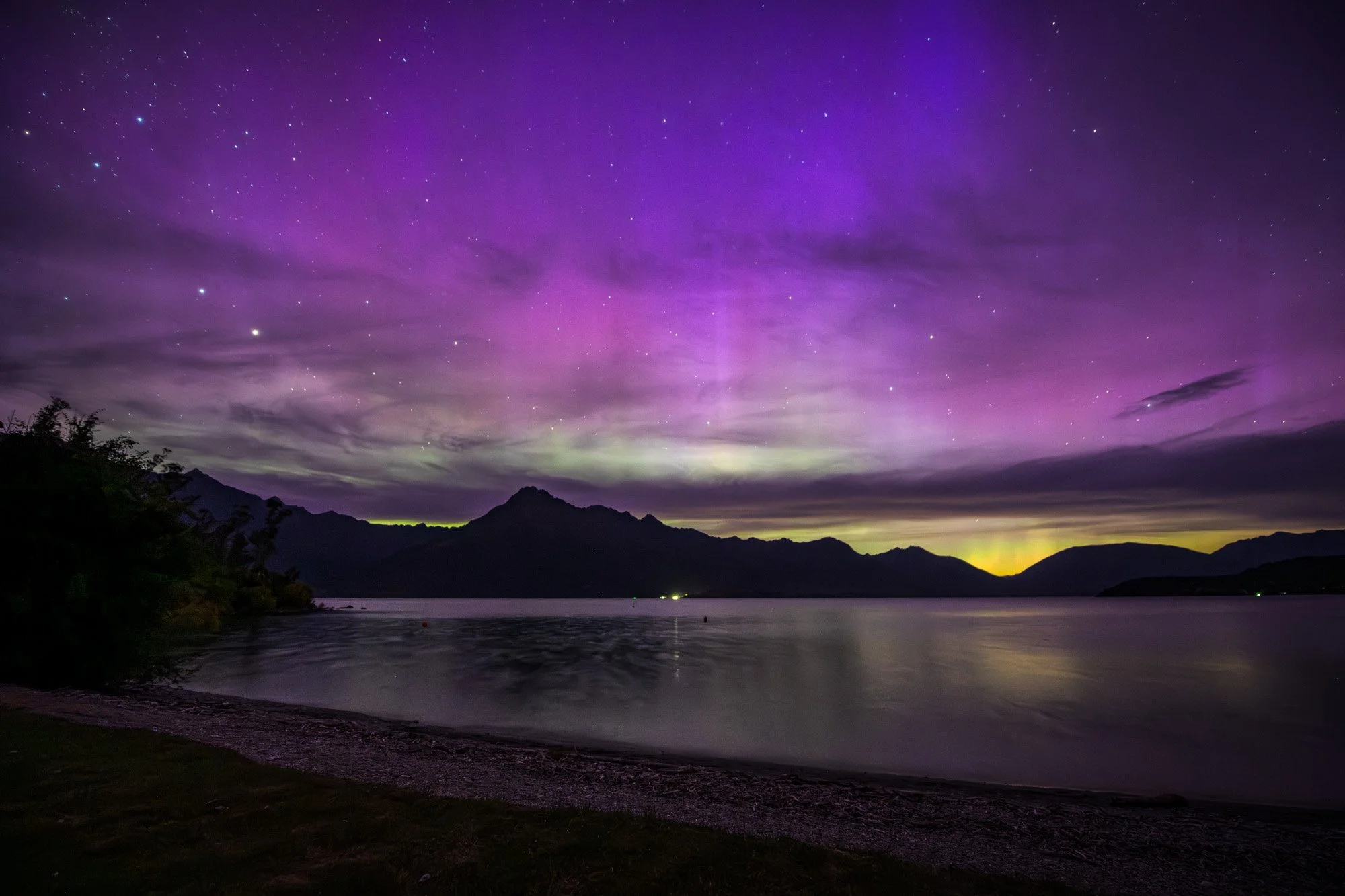 Magenta and white aurora beams rising vertically from behind dark mountain silhouettes against a twilight sky over Lake Wakatipu