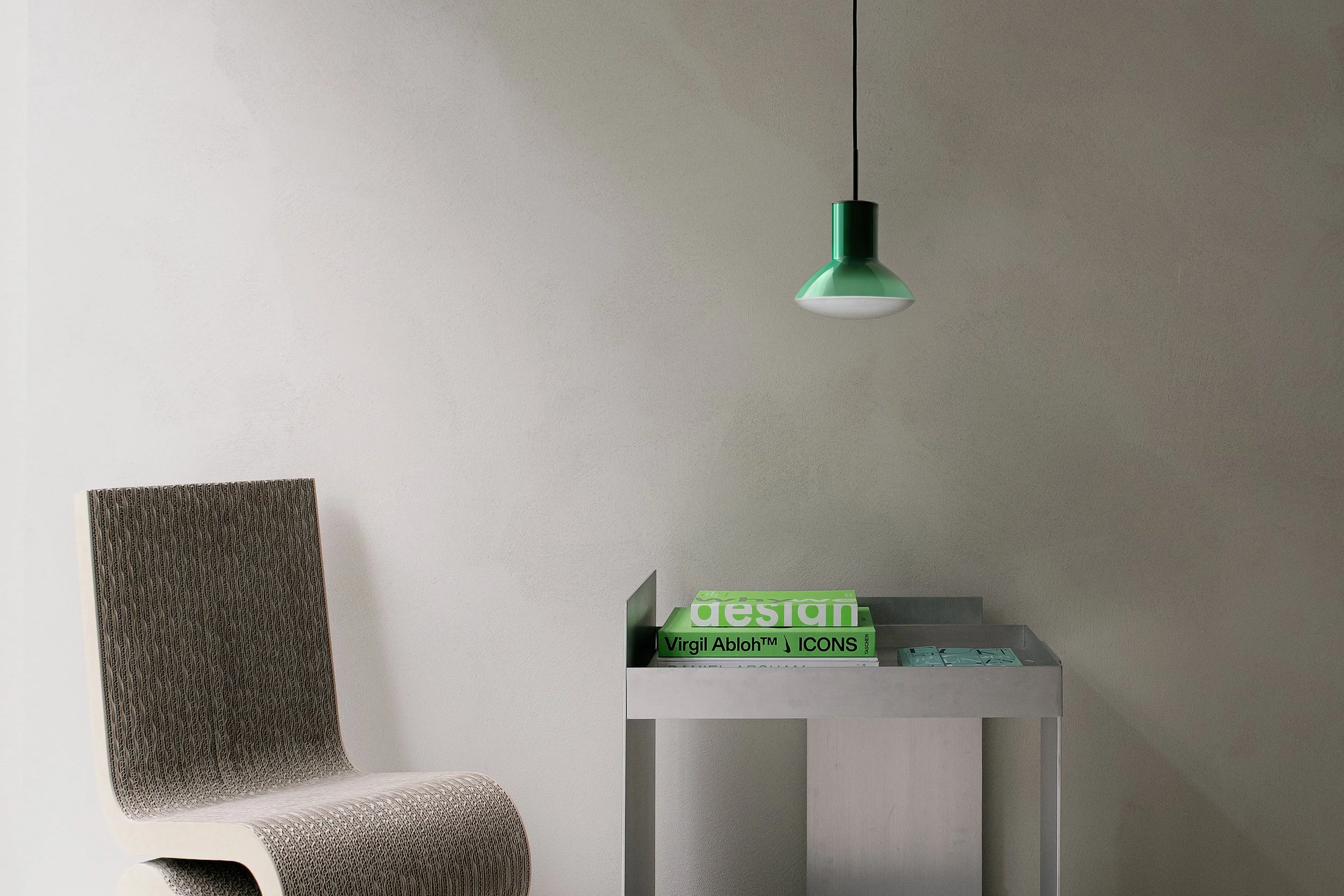 Ein moderner Innenraum mit einem grauen Stuhl, einen weißen Beistelltisch mit designbezogenen Büchern und eine grüne Deckenlampe.