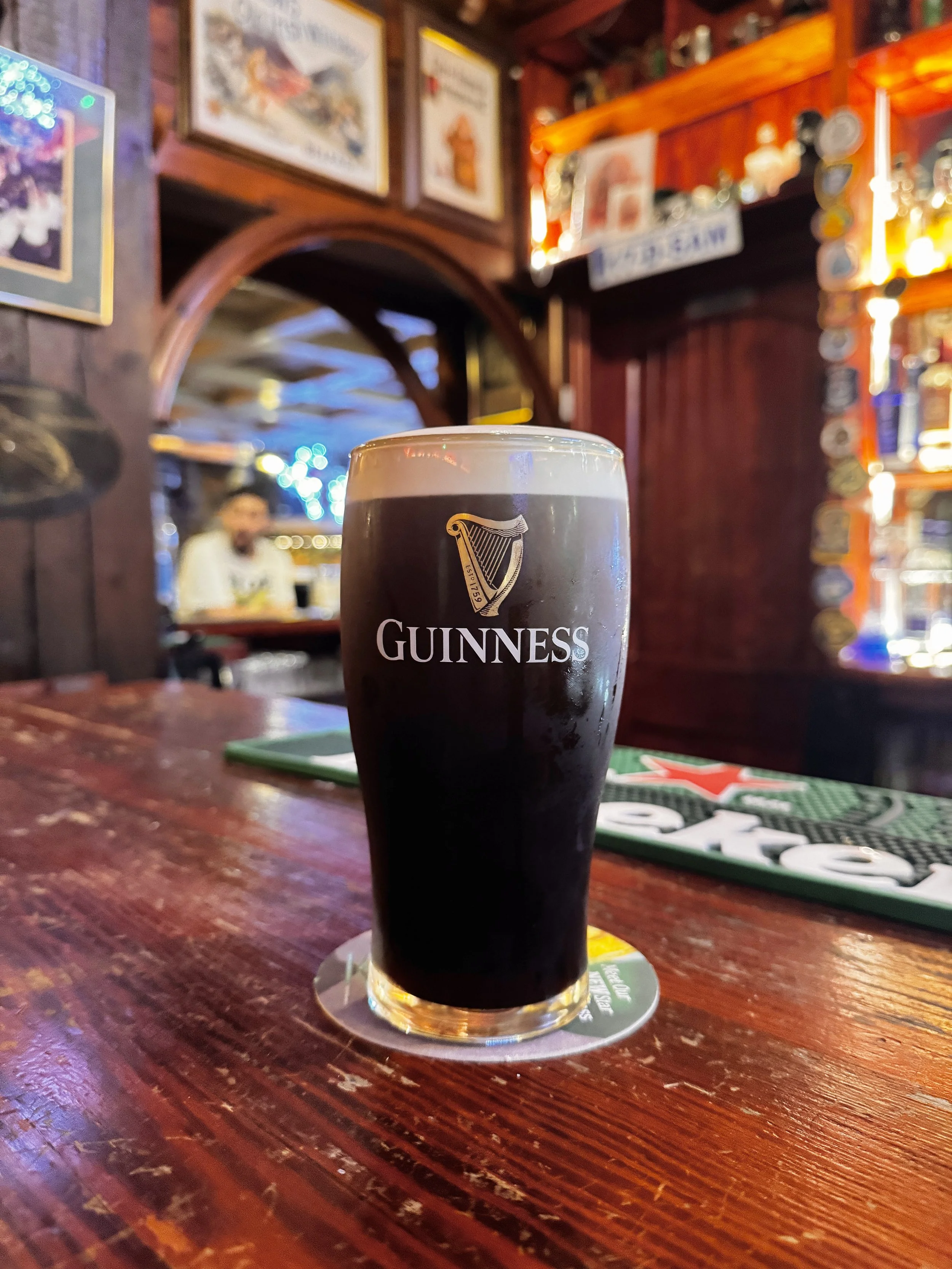 A pint of Guinness beer on a wooden bar counter inside a pub, with a background of blurry bar decor and a person sitting at the bar.
