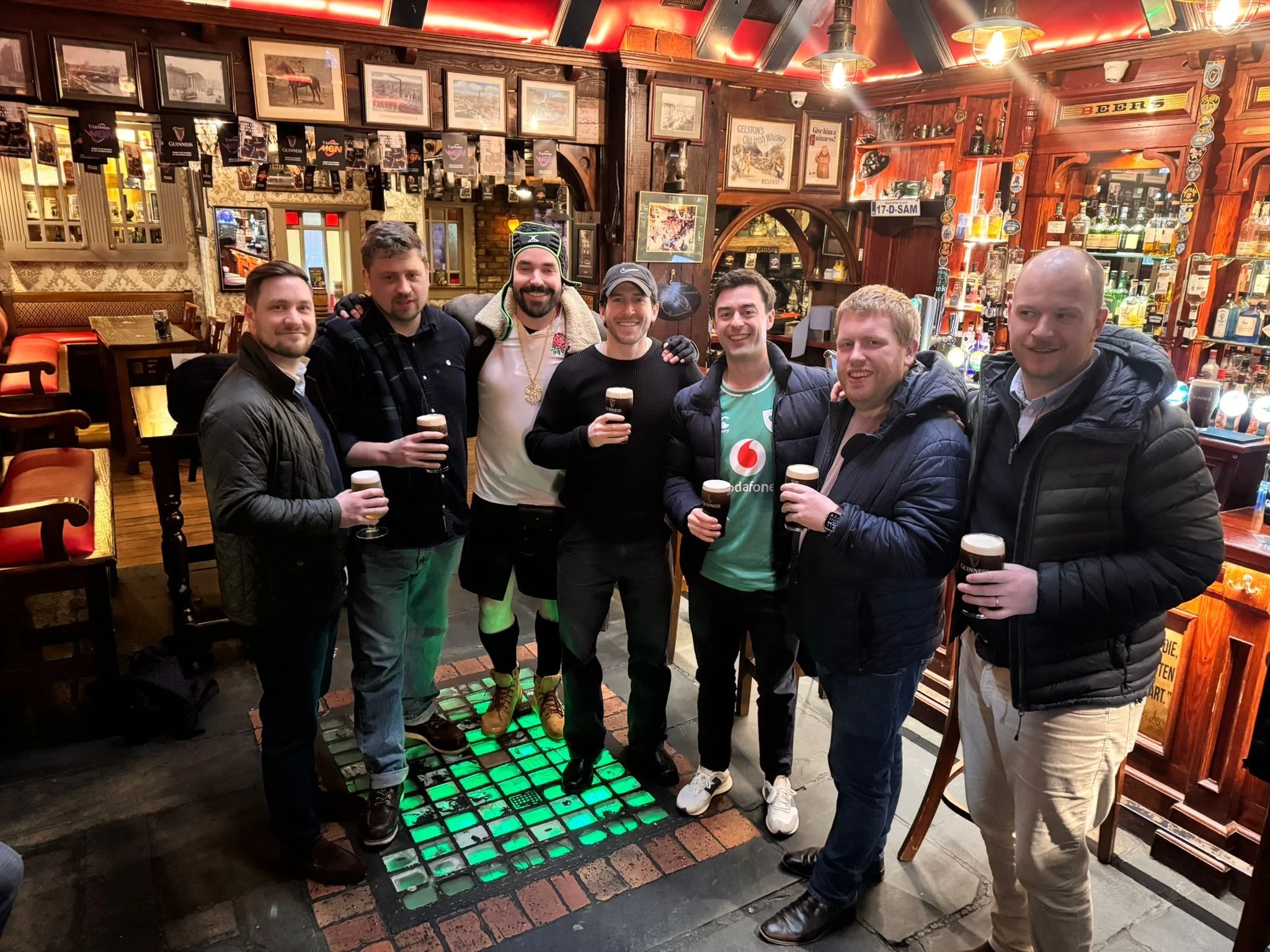 A group of seven men standing inside a pub, smiling and holding pints of dark beer. The pub has a cozy, traditional decor with wooden panels, framed pictures, and various bottles of alcohol behind the bar. The floor features a digital dance floor wit