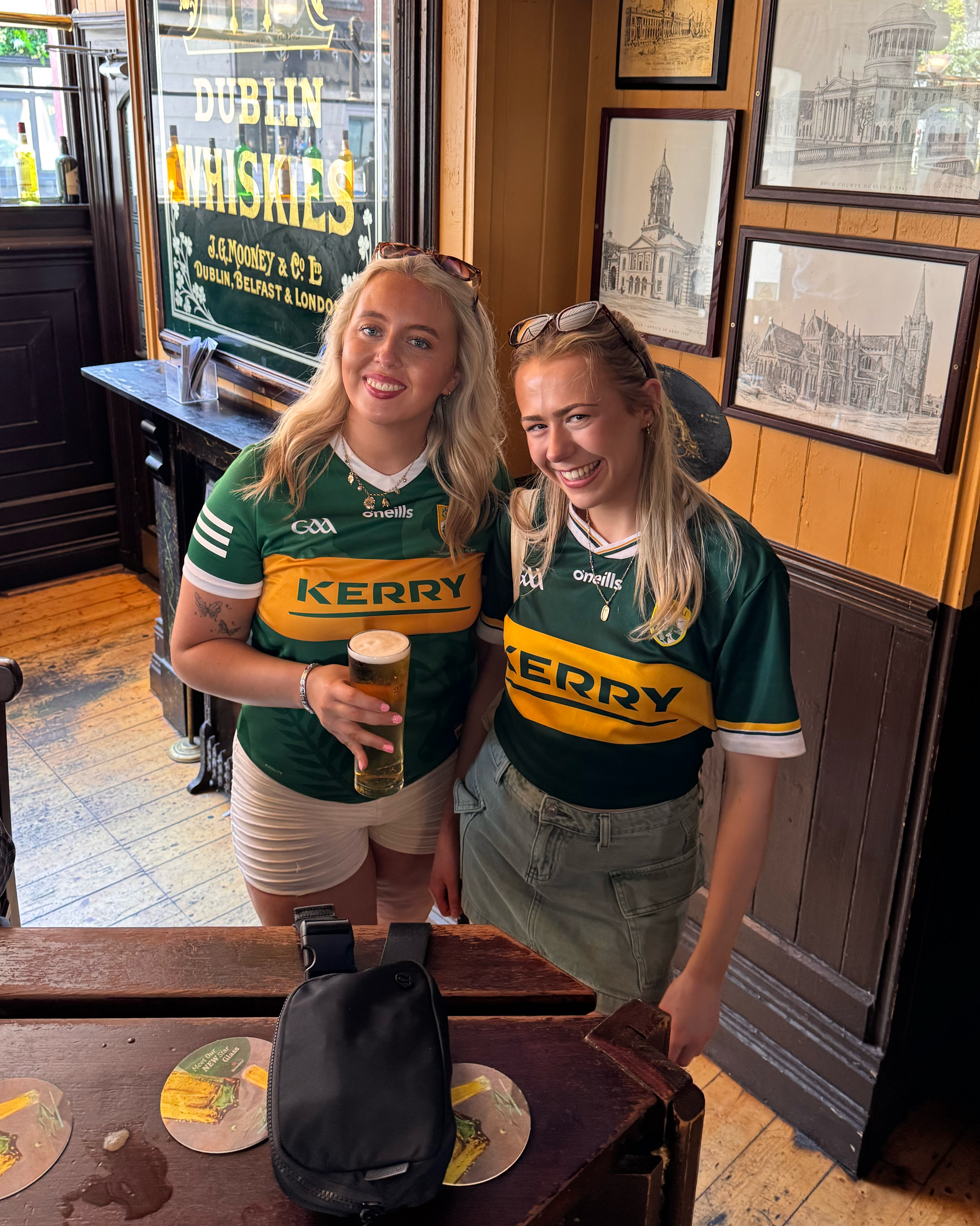 Two young women wearing green and yellow Kerry football jerseys are smiling at the camera inside a pub or restaurant. One is holding a glass of beer. The background features framed sketches of Irish landmarks on a yellow wooden wall and a large windo