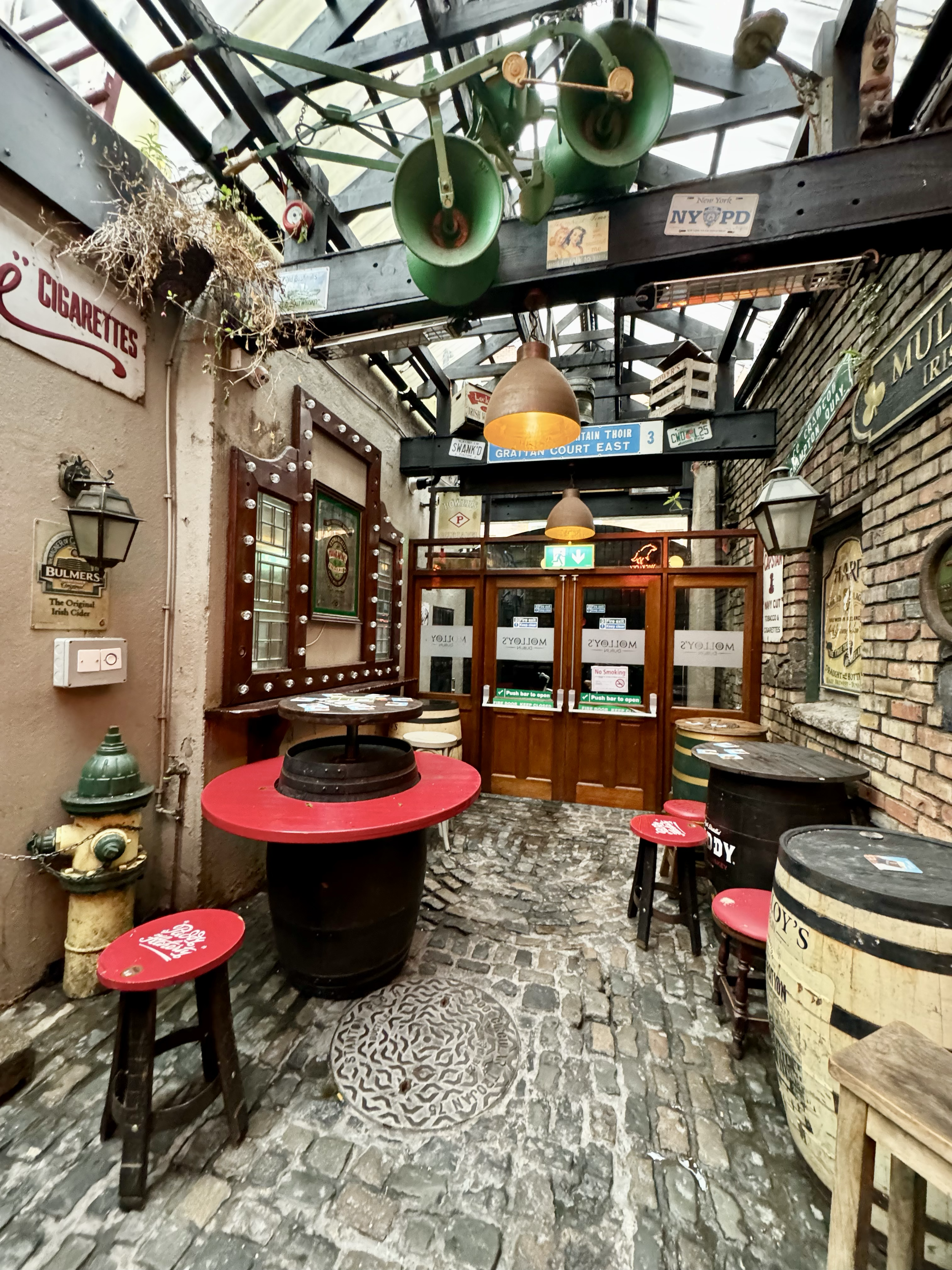 A rustic outdoor patio with cobblestone flooring, decorated with vintage beer barrels, small tables, and stools with red seats featuring white logos. The space is enclosed by brick and plaster walls and includes hanging lamps, old signs, and plants o