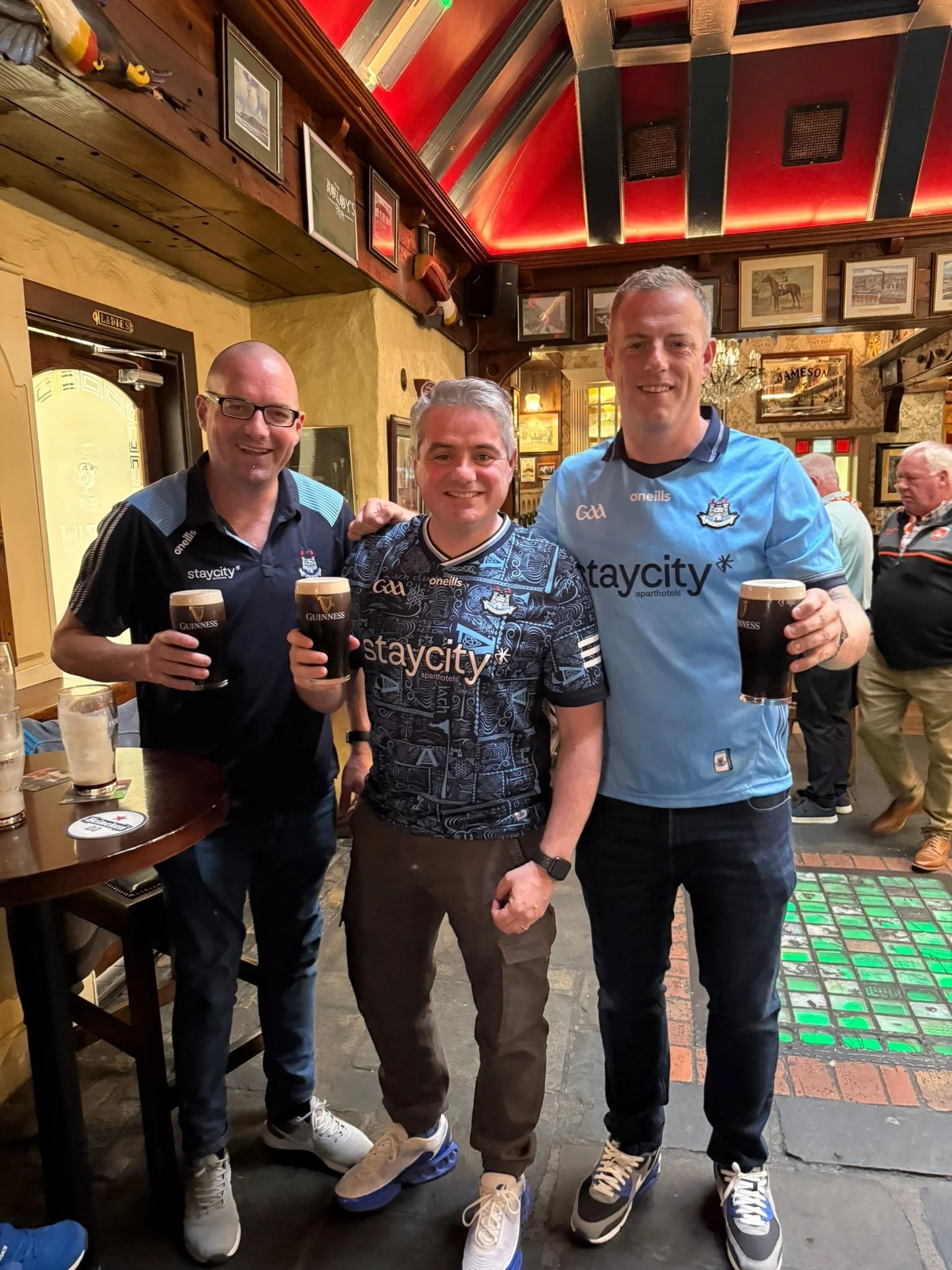 Three men holding Guinness cups and smiling, standing in a pub with wooden walls, framed pictures, and decorative lighting.