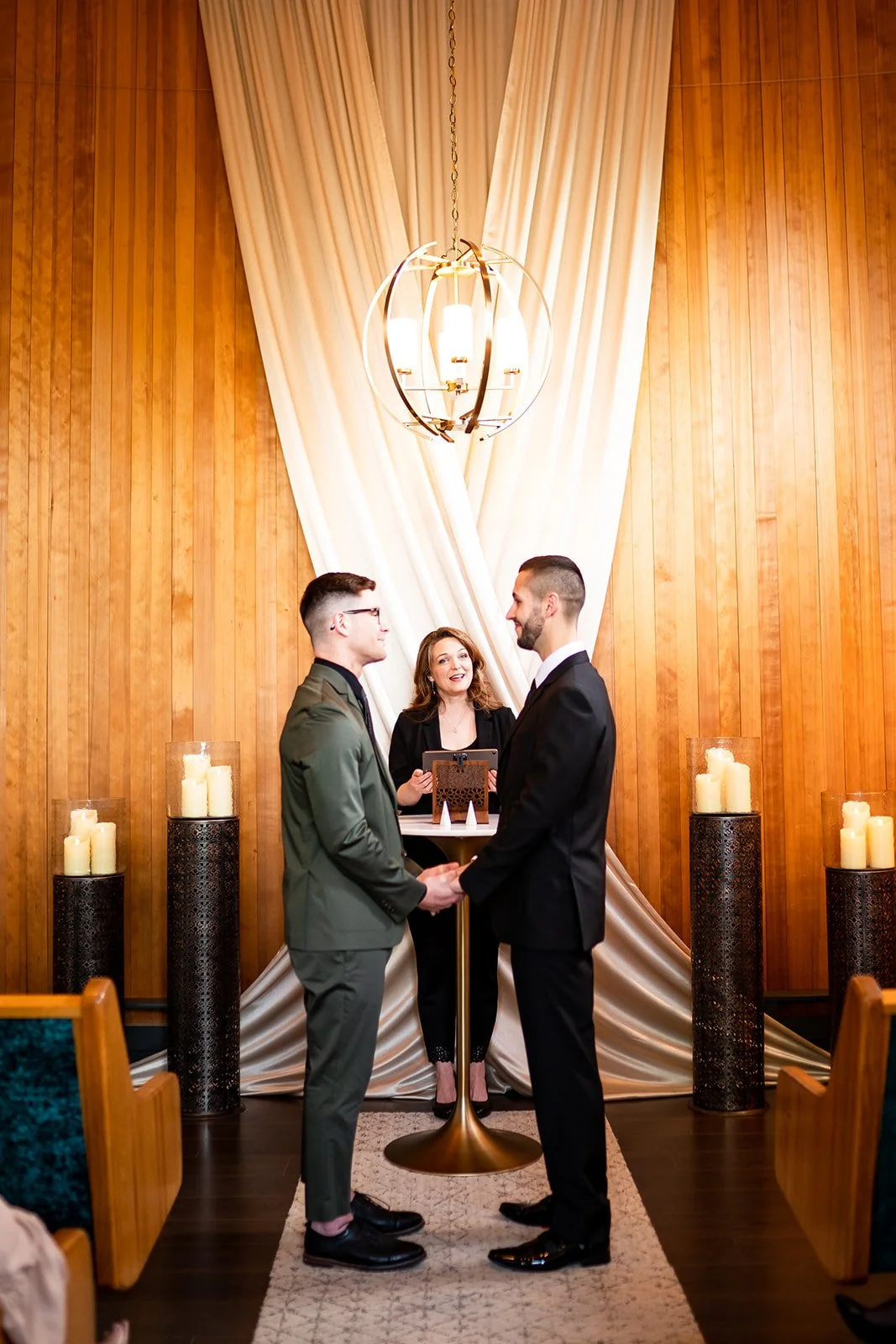 A wedding ceremony with two men holding hands, facing each other, and officiant smiling behind a small table, in a Mid Century Modern wooden-paneled room with candles and draped white fabric.