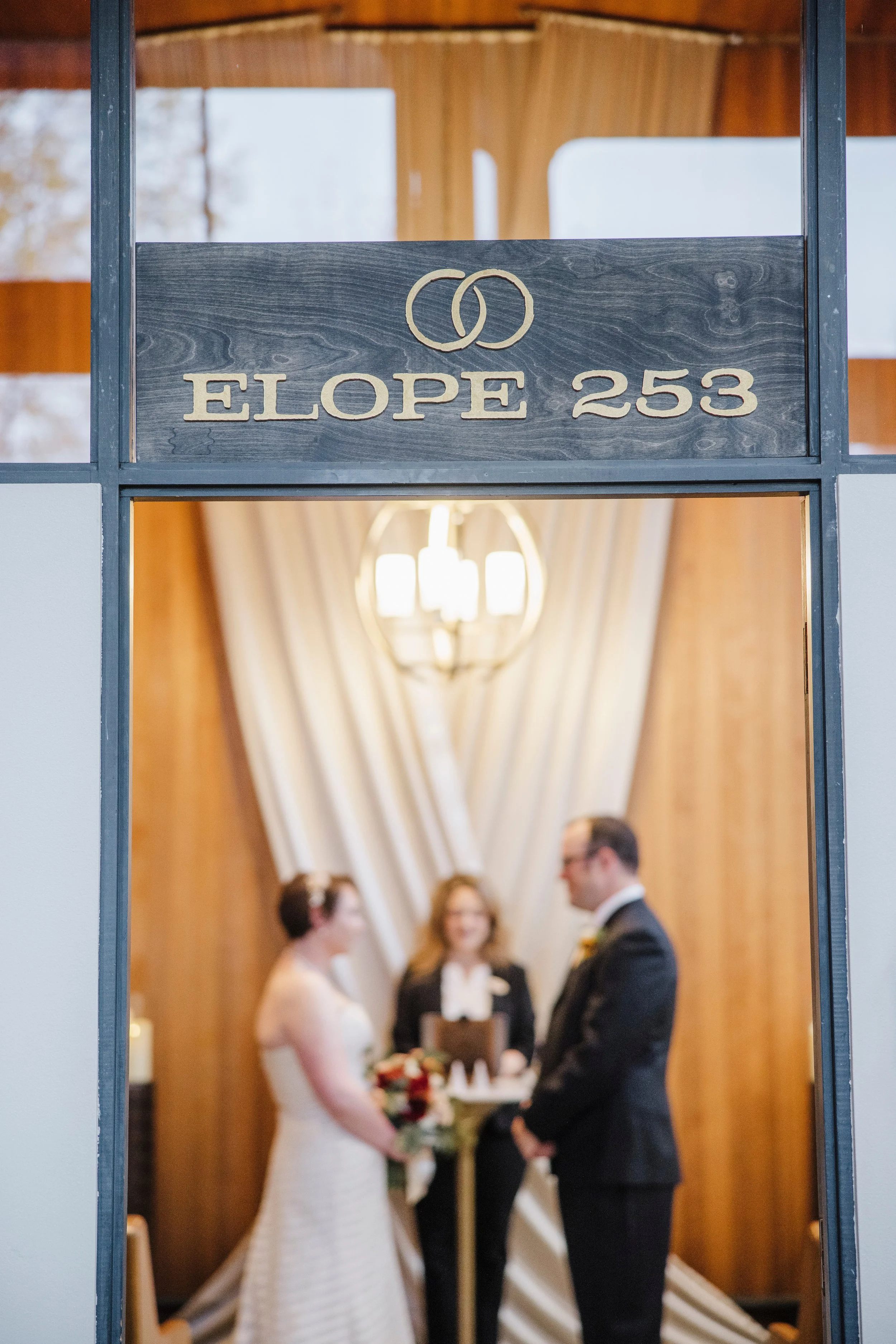 A couple getting married in a small indoor ceremony, with a minister officiating. The focus is on the couple, with the bride in a white dress and the groom in a dark suit. The background shows warm wooden walls and a chandelier, with friends and fami