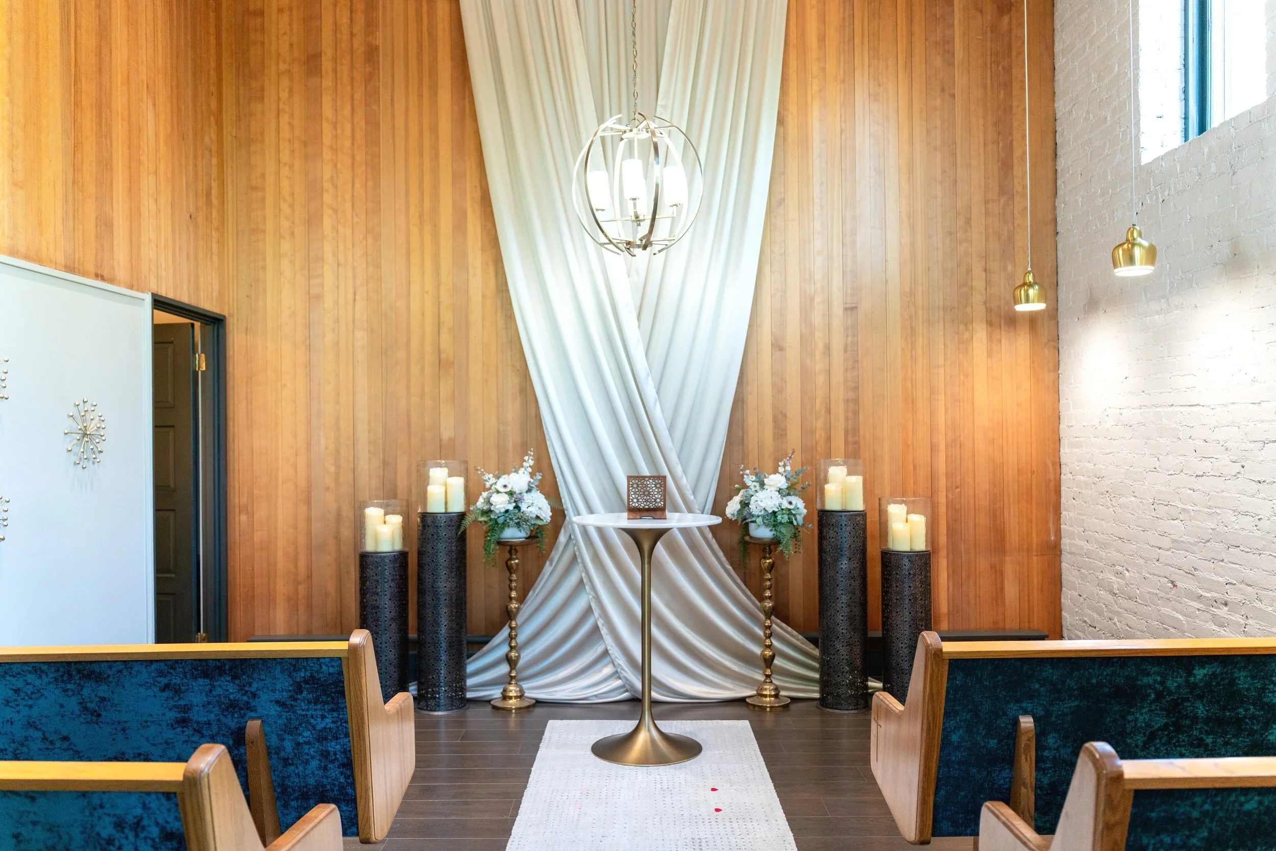 Interior of a church or chapel setup for a wedding with a draped white cloth backdrop, flowers, candles, and a small table at the front, with pews on either side.