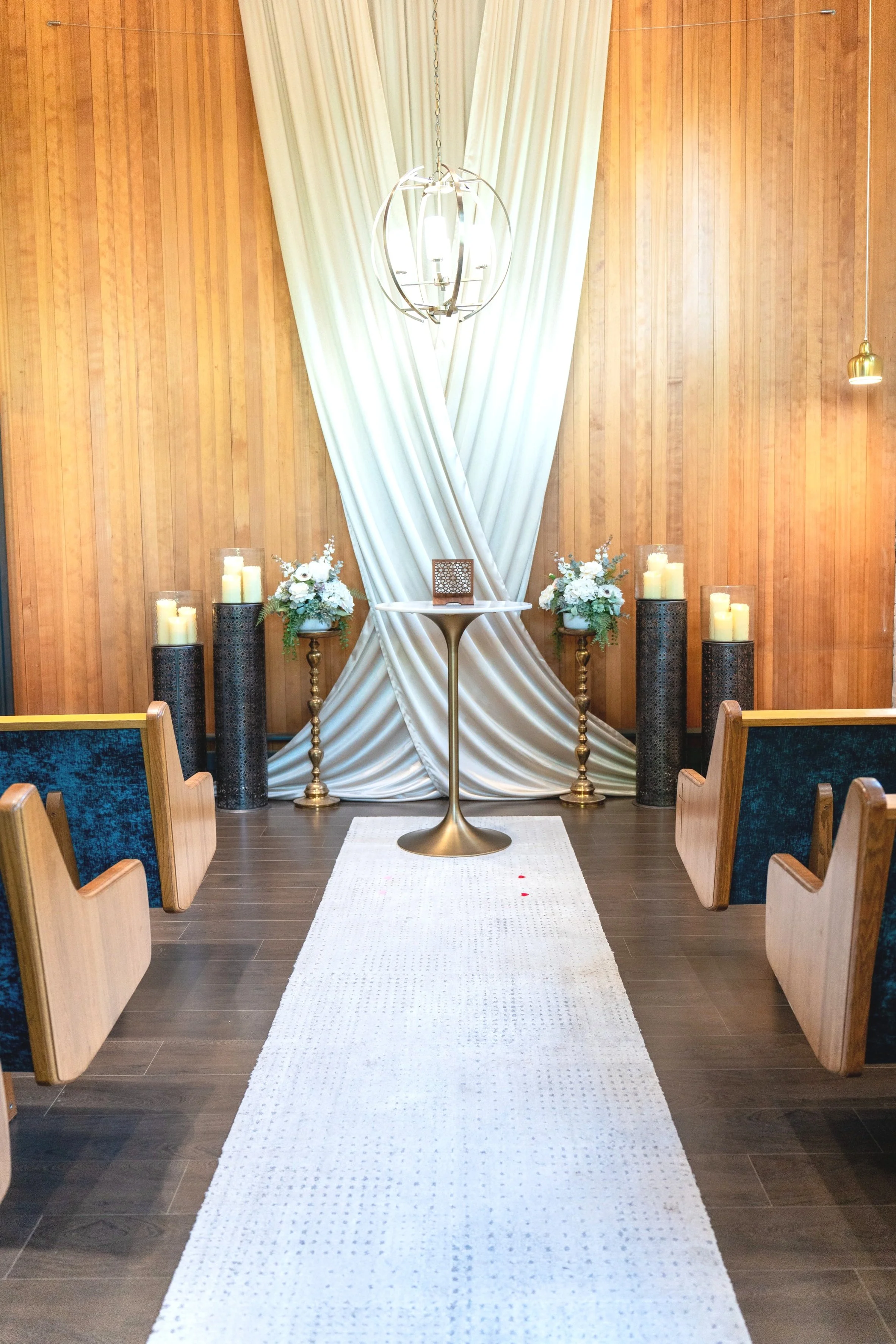 A church interior with wooden walls, a white draped altar, candle holders, floral arrangements, and chairs facing the altar.