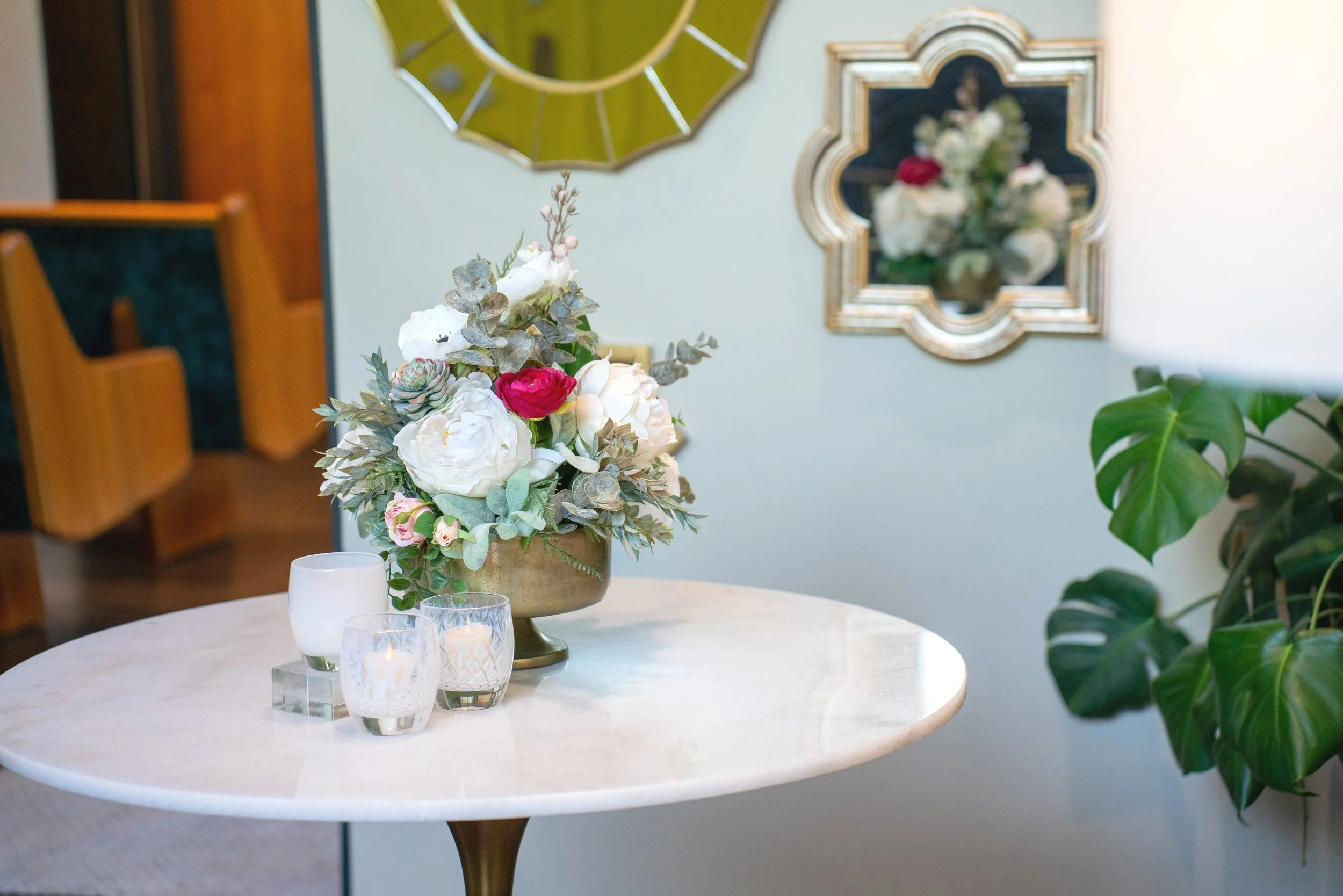 A floral arrangement on a white marble round table, with candles, in a room with wall mirrors and a wooden chair.