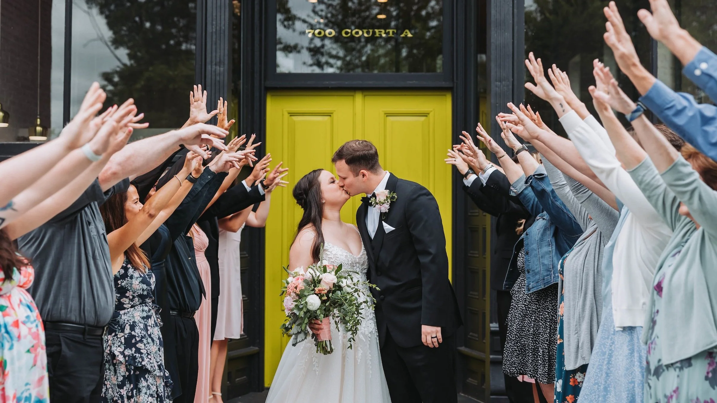 A bride and groom kissing in front of a crowd with arms raised, gathered outside a building with a bright yellow door and black frame, marked with the address '700 Court A'.