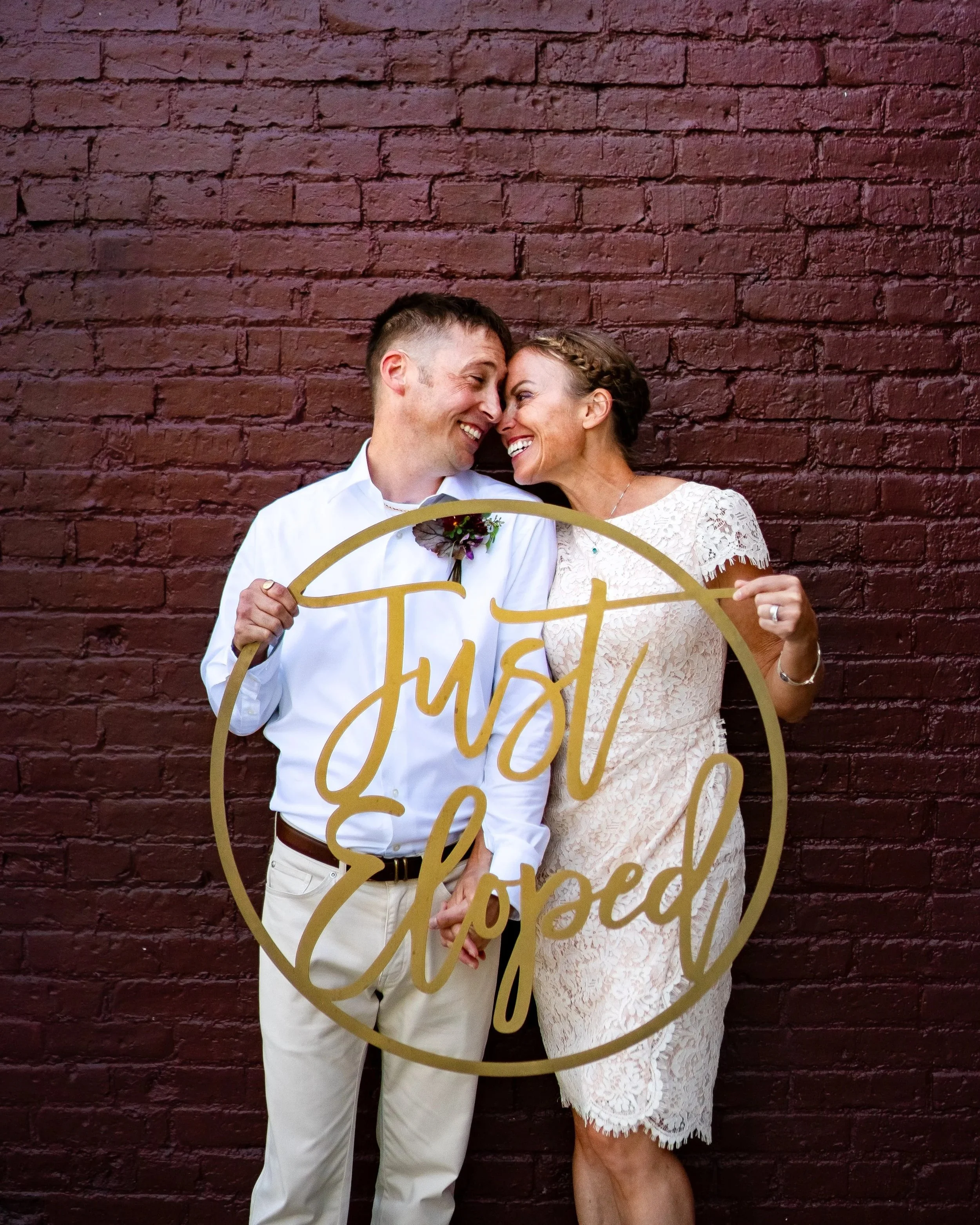 A couple is smiling and touching foreheads, holding a gold 'Just Married' sign in front of a red brick wall.
