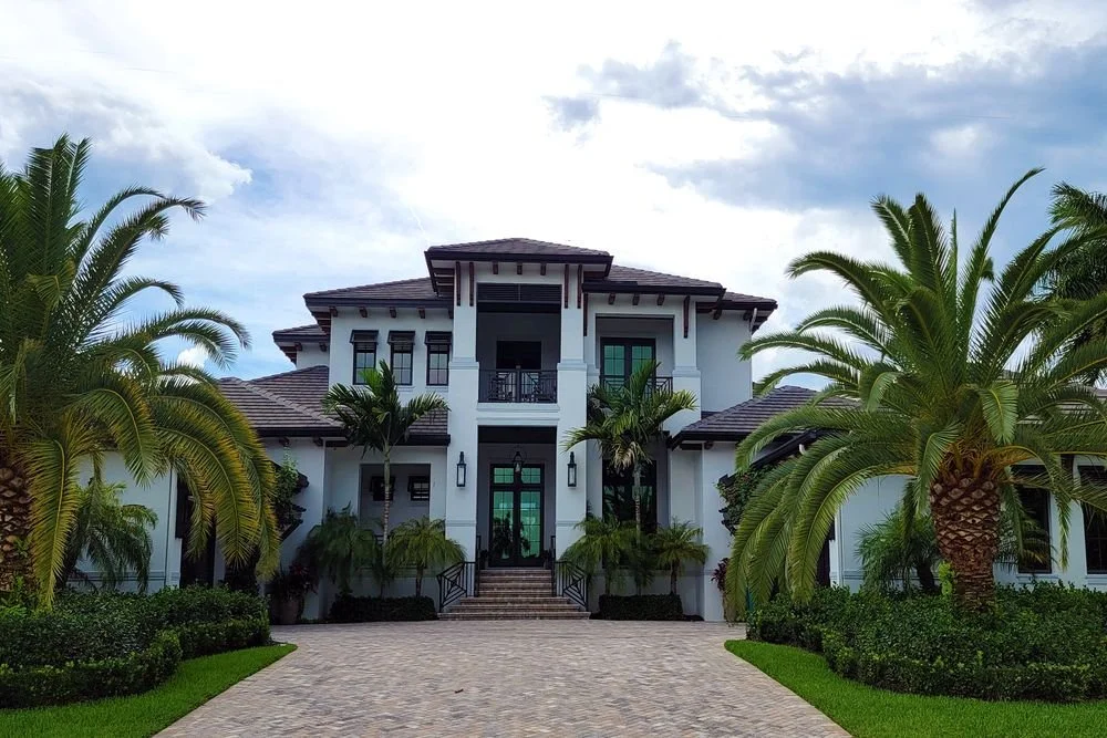 Large two-story white home with a light-colored paver driveway, palm trees, and manicured hedges