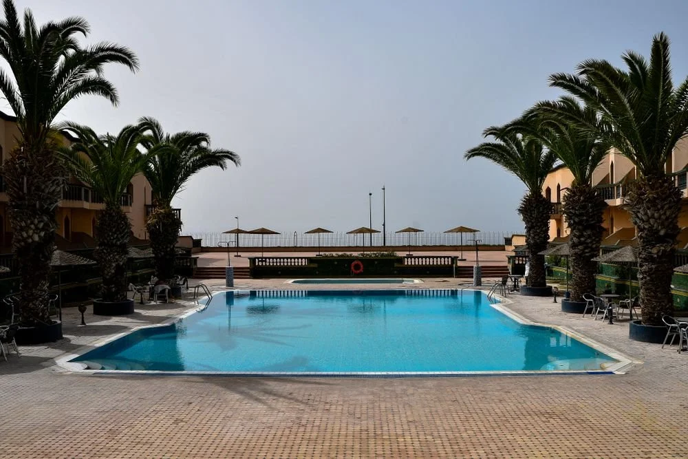 Outdoor swimming pool surrounded by a light-colored paver deck, palm trees, lounge chairs, and ocean views