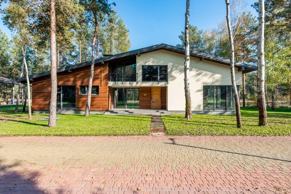 Modern two-story house surrounded by tall trees, with a spacious brick paver driveway and green lawn