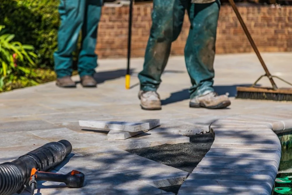 Workers installing gray stone patio pavers with landscaping tools and a broom nearby
