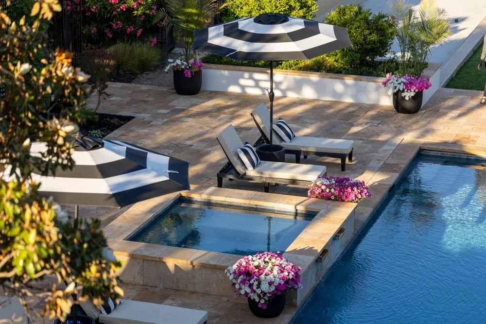 Tan stone paver pool deck with a raised spa, lounge chairs, striped umbrellas, and colorful flowers