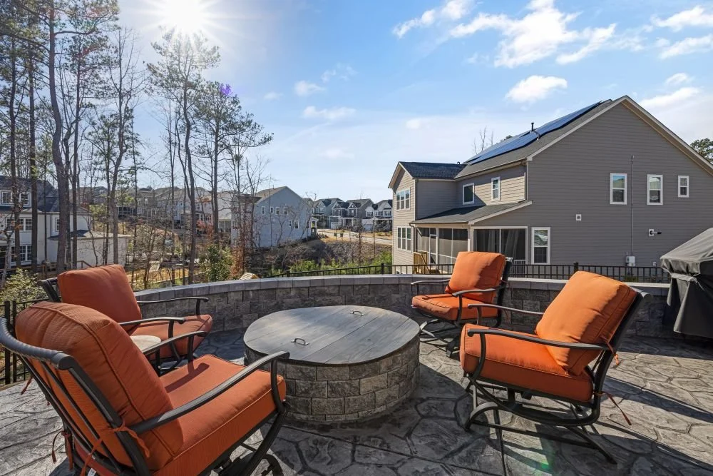 Stone paver patio with a round fire pit, orange cushioned chairs, and a low retaining wall