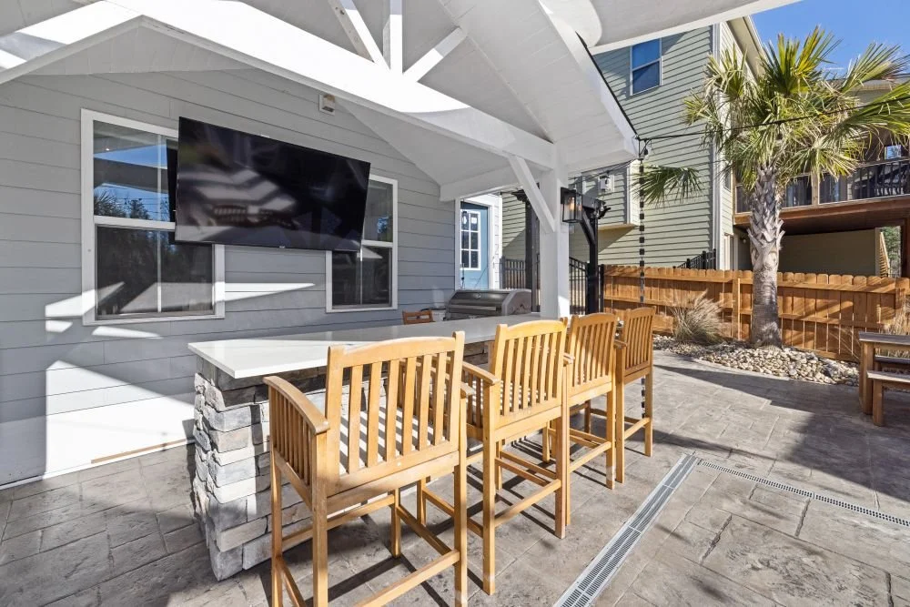 Covered outdoor patio with gray stone pavers, a built-in bar, wooden stools, wall-mounted TV, and palm tree