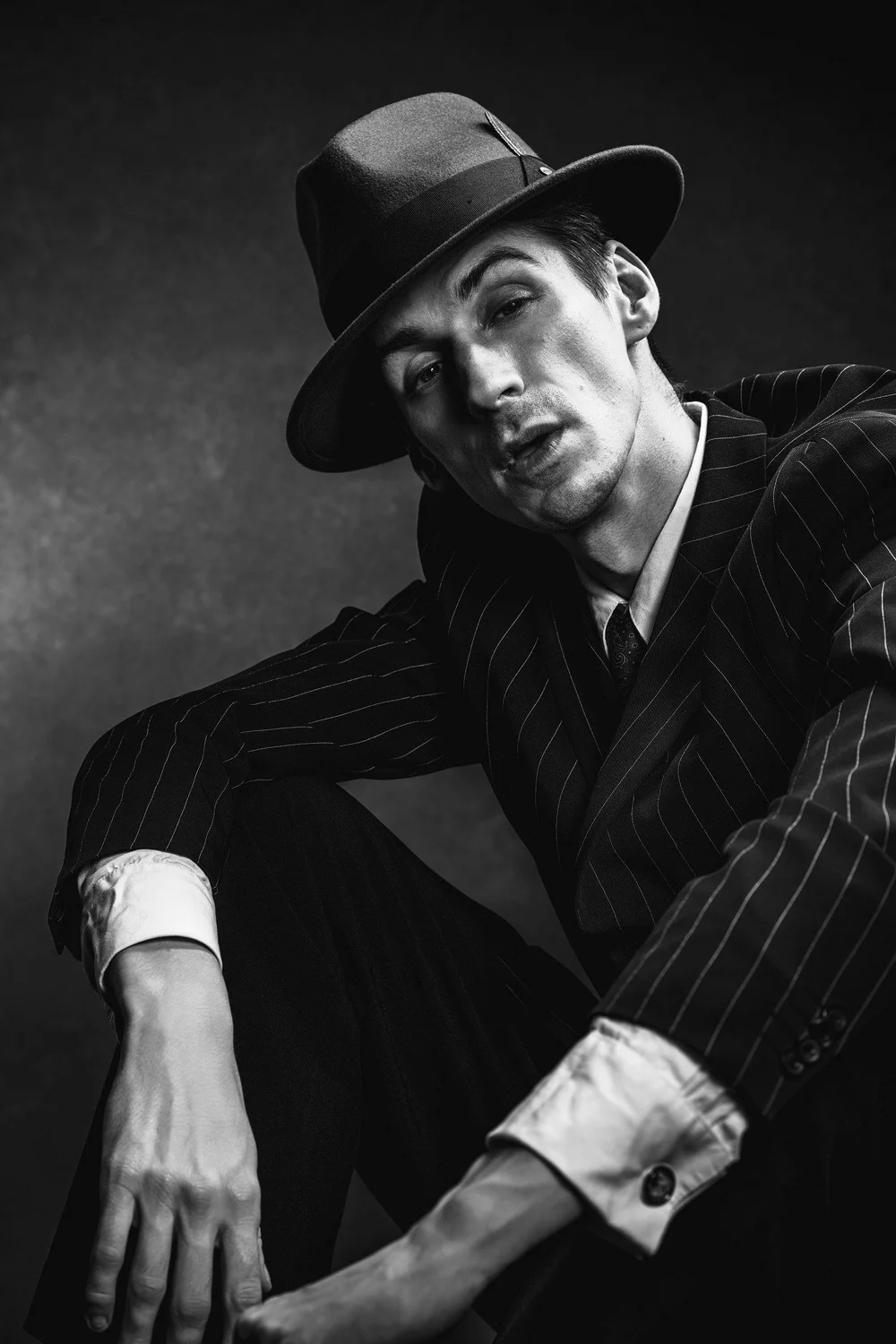 Black-and-white portrait of a seated man in a pinstripe suit and fedora, leaning forward.