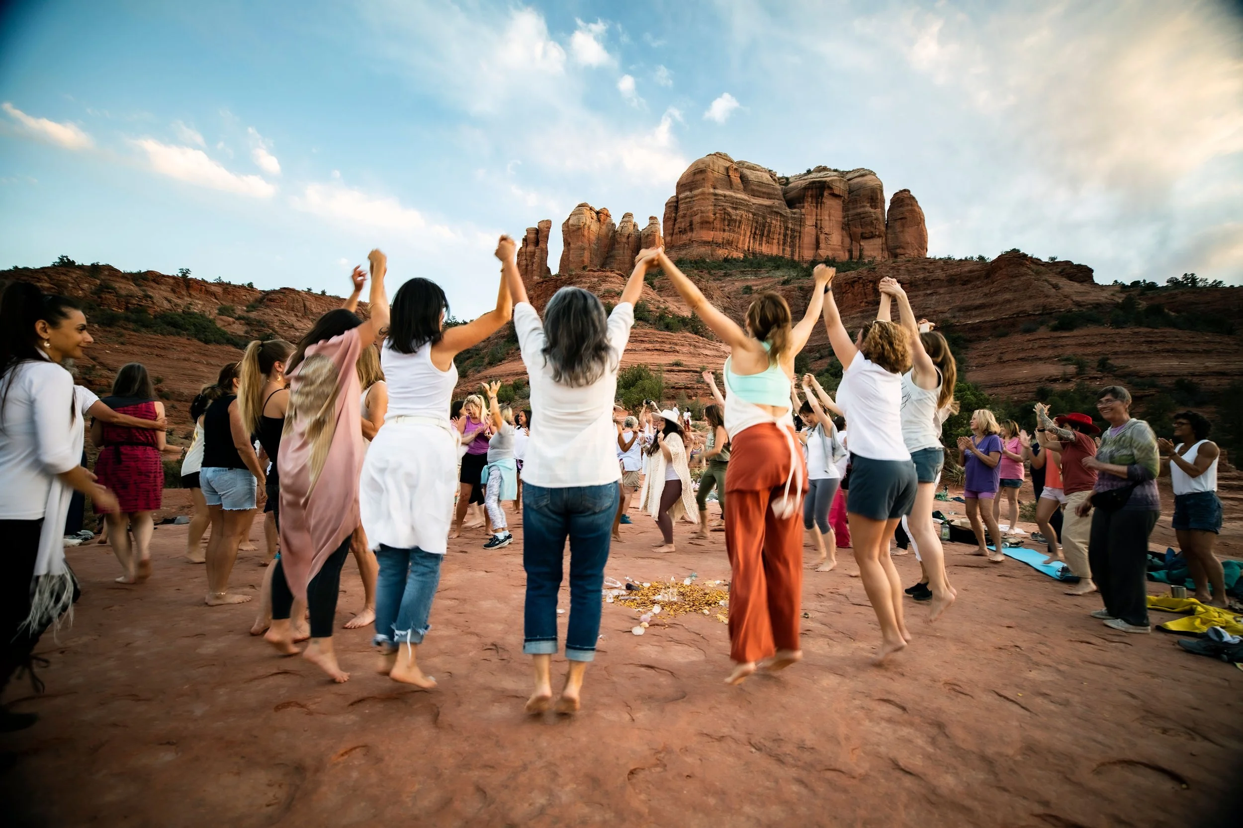 A group of people participate in a yoga session outdoors on a red rock surface with a desert mountain landscape in the background, under a partly cloudy sky.